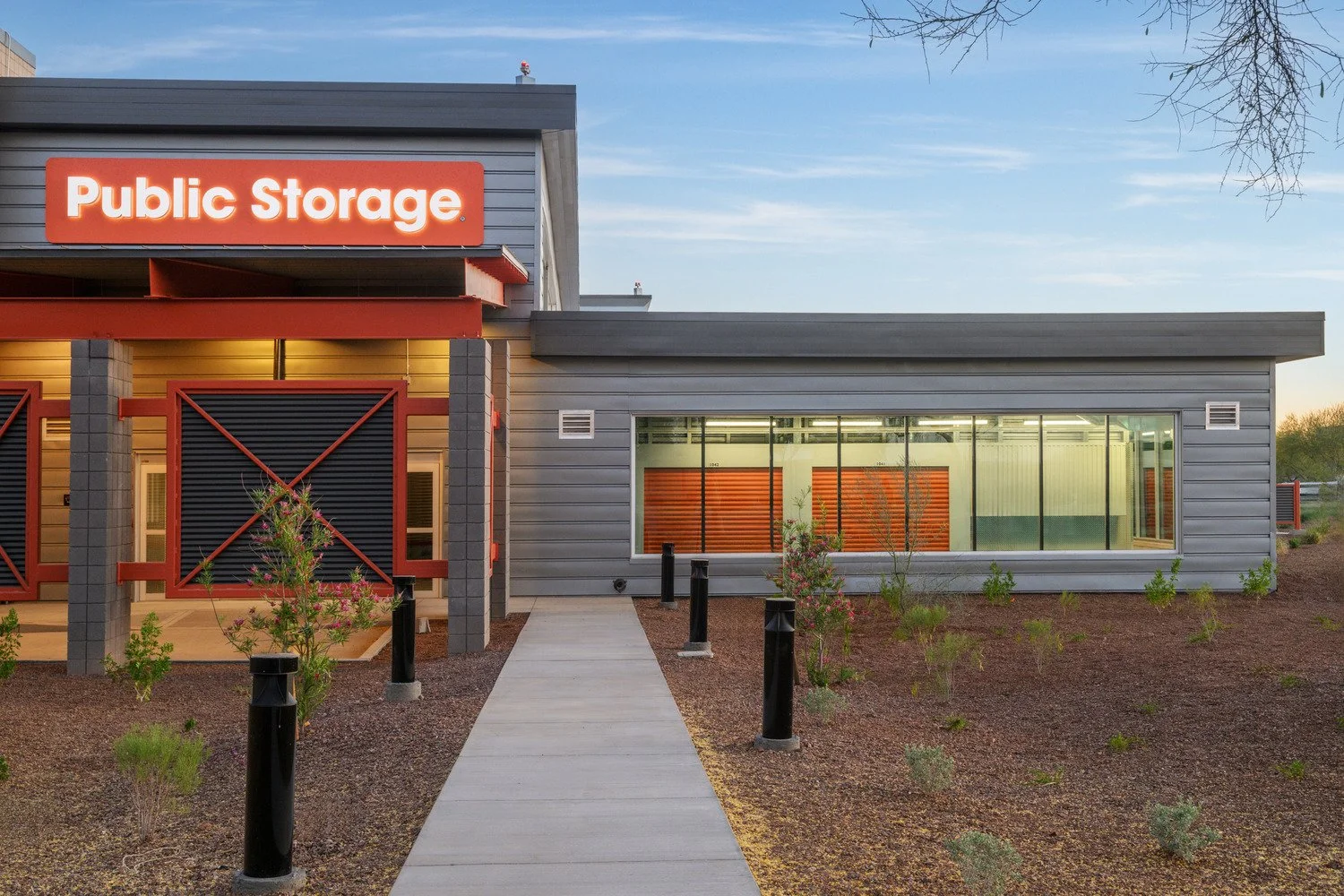 Exterior view of a public storage facility with a sign reading 'Public Storage.' The building has gray siding, large glass windows, black bollards along the walkway, small plants, and a clear sky in the background.
