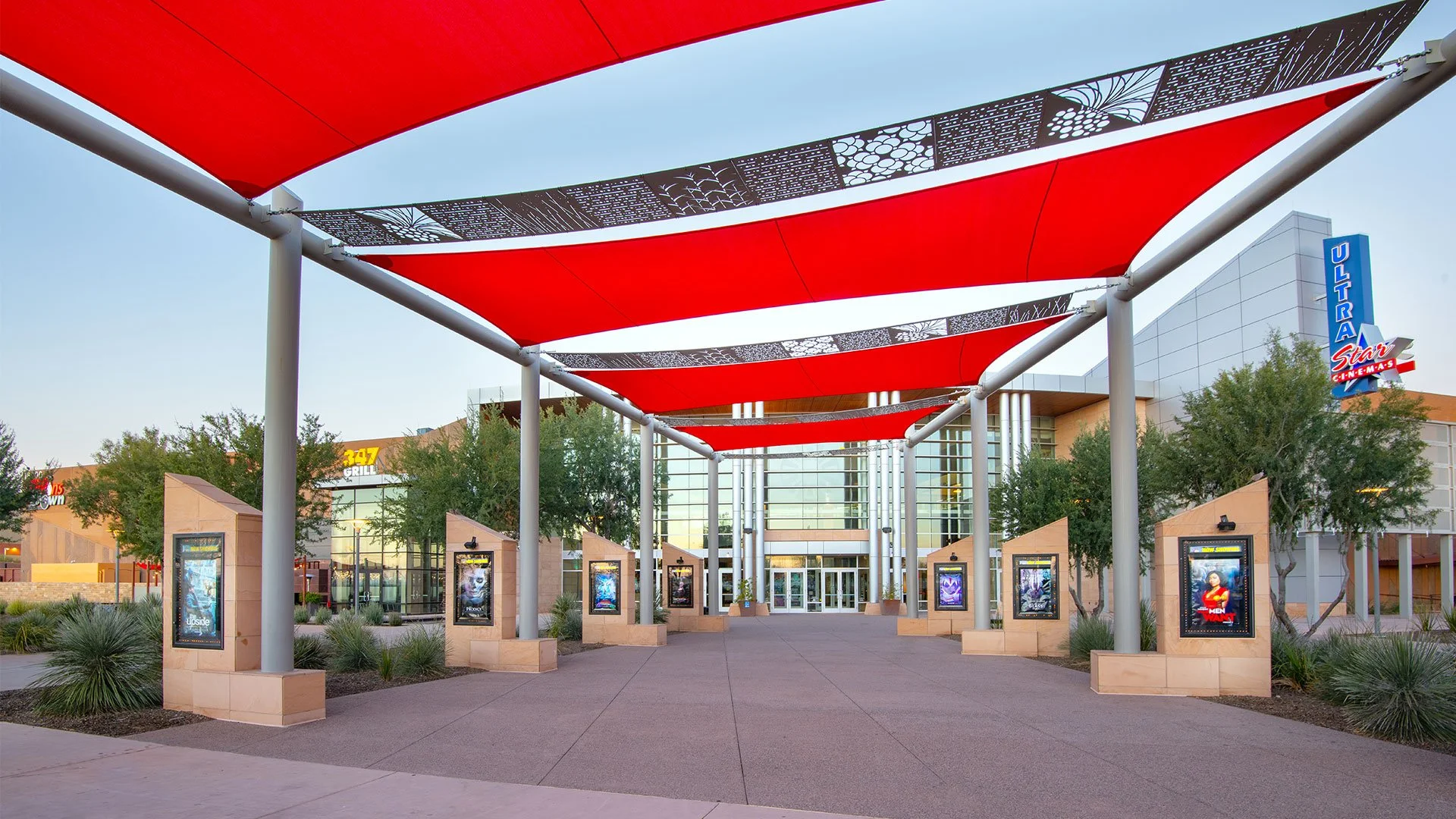 An outdoor shopping mall entrance with red canopy shades, movie posters along the walkway, and a modern glass building in the background.