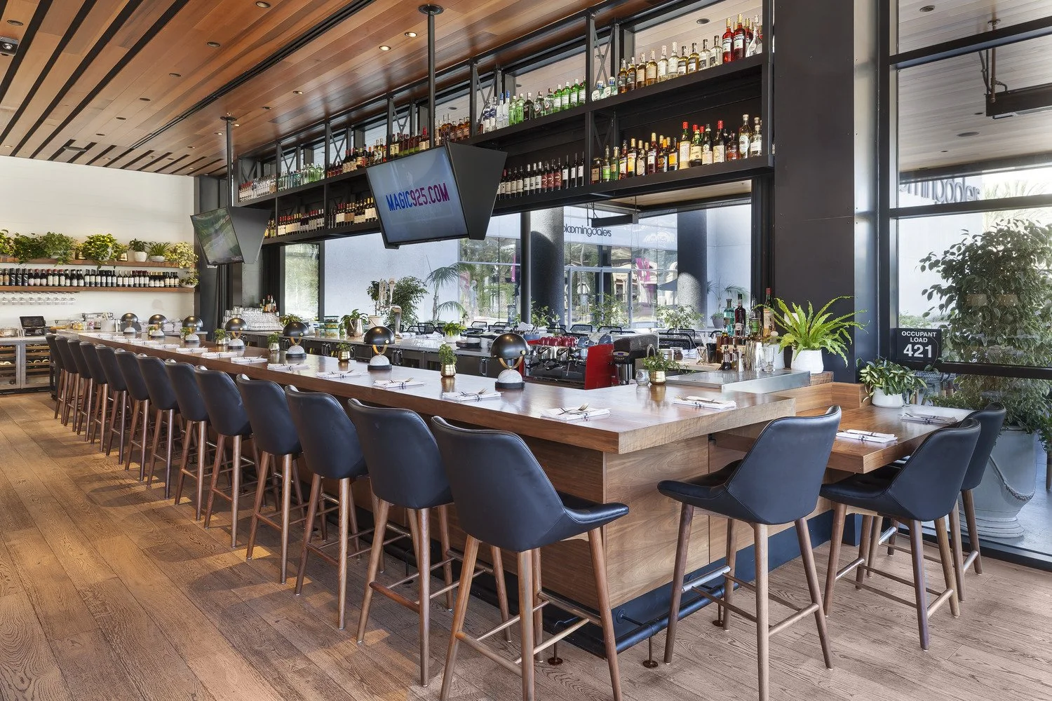 Modern restaurant bar with a long wooden counter, black chairs, shelves of liquor bottles, and large windows with plants outside.