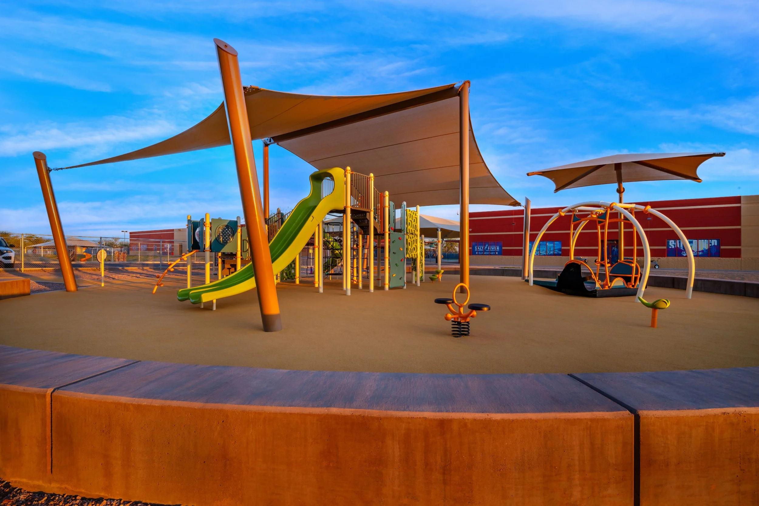An empty outdoor playground with a slide, climbing structures, shade canopies, and a sandbox area, set against a blue sky with clouds.