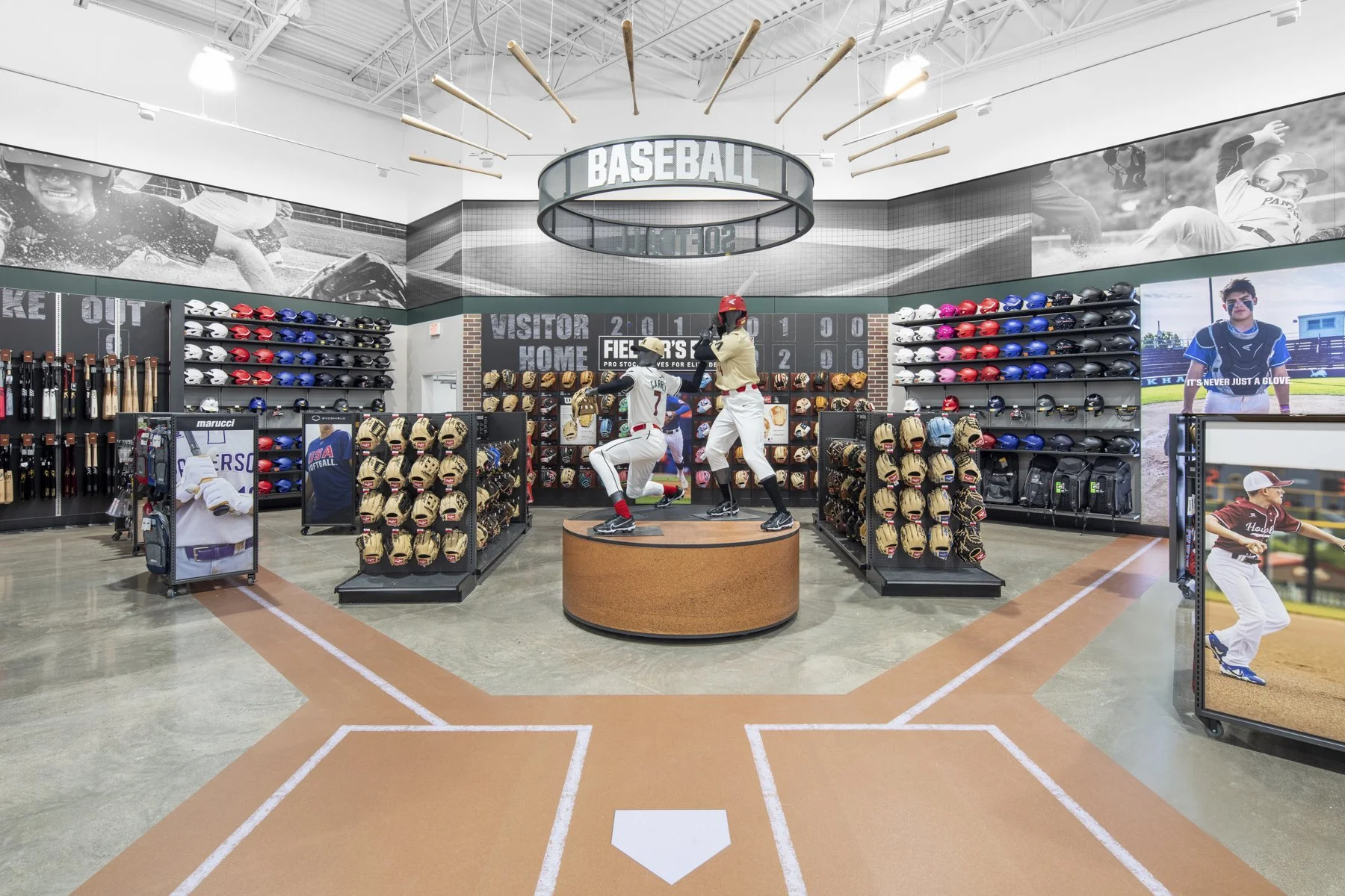 Indoor baseball store with helmets, bats, and baseball gloves on display. Mannequins wearing baseball uniforms and helmets on a circular platform. Background features baseball-themed murals and a scoreboard.