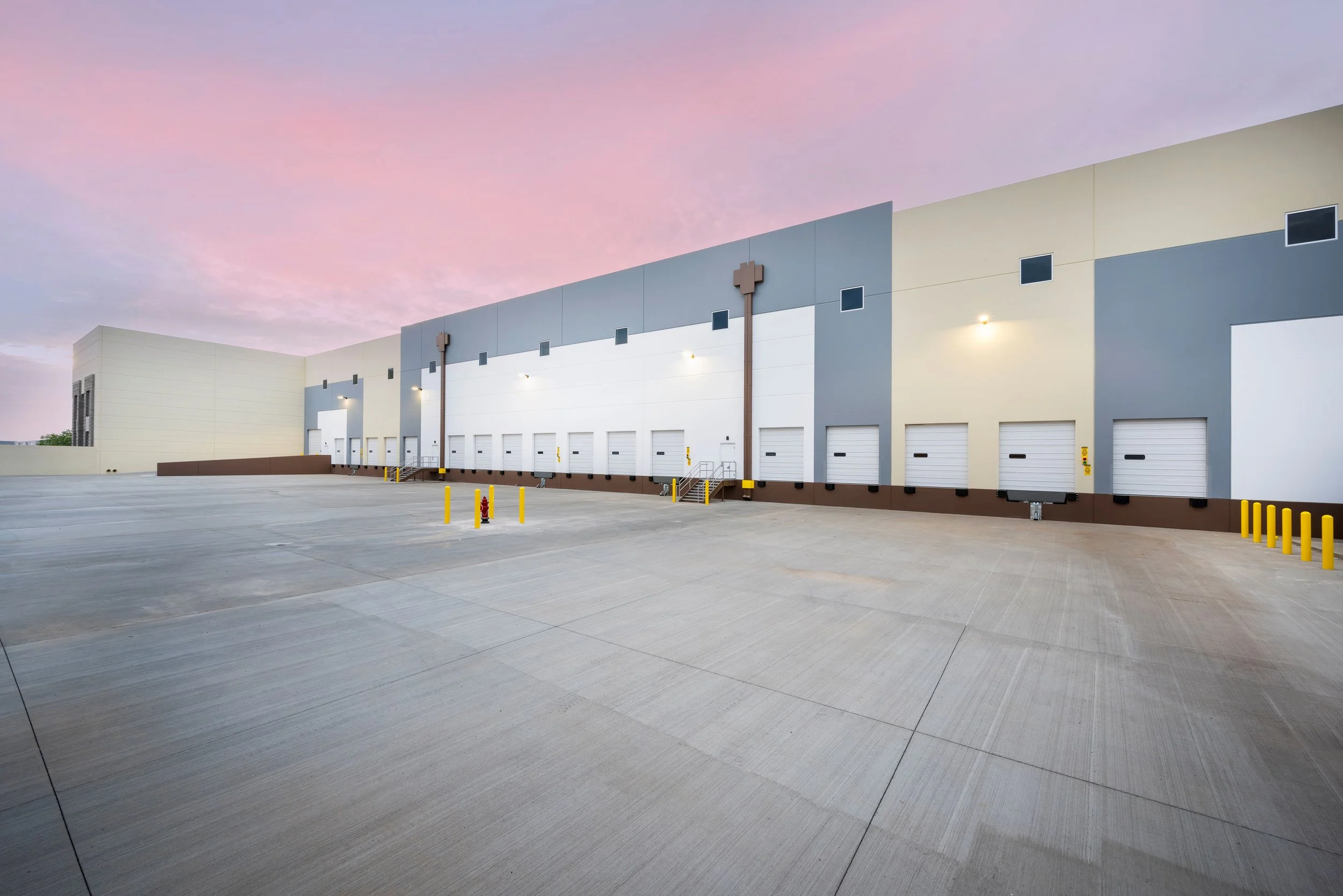 Empty warehouse loading dock with multiple bays and closed white roll-up doors, concrete pavement, yellow safety bollards, modern industrial building exterior at sunset.