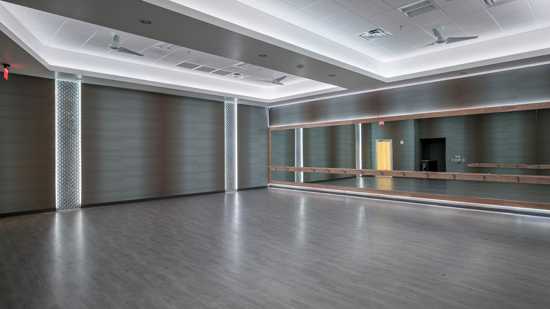 Empty dance studio with wooden floors, a large wall mirror, ballet barres, and ceiling fans.