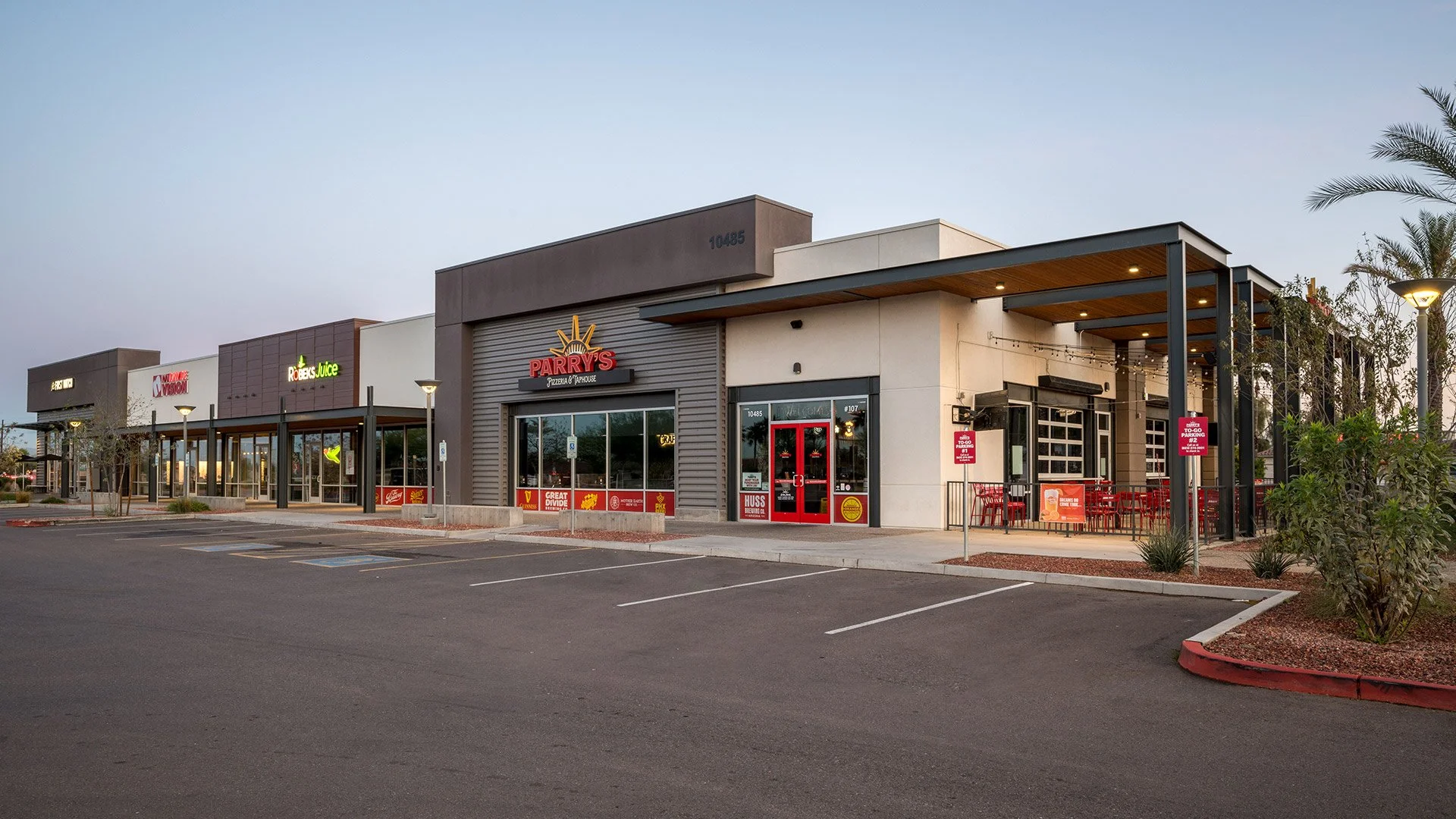 Empty parking lot in front of a shopping plaza with several retail stores including Parry's Pizza and juice bar, with some small trees and bushes, under a clear sky.