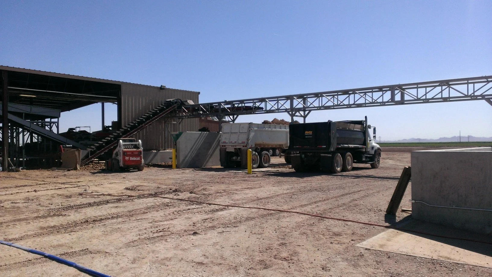 Construction site with trucks, concrete barriers, and an elevated conveyor system under clear blue sky.