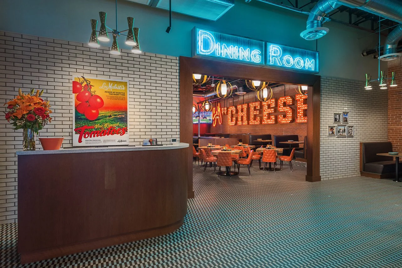 Interior of a restaurant with a reception desk, a neon sign reading 'Dining Room', and a cozy seating area with tables and chairs. The decor includes brick walls, vintage-style posters, and modern lighting fixtures.