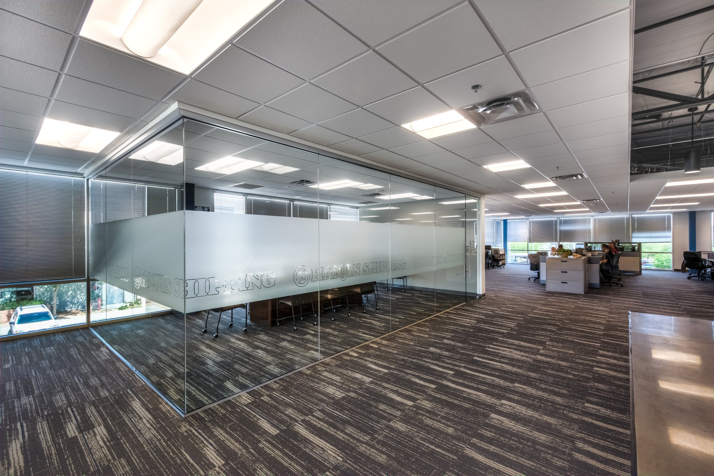 Modern office space with glass-walled conference room, partial view of open workspace with desks and chairs, large windows with blinds, ceiling with recessed lighting, carpeted floor.