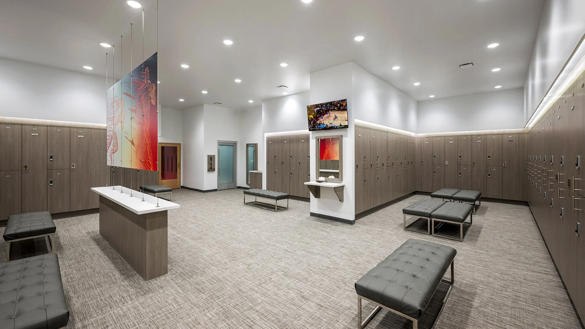 Empty locker room with multiple benches, lockers lining the walls, a TV on the wall, a mirror, and a decorative banner hanging from the ceiling.