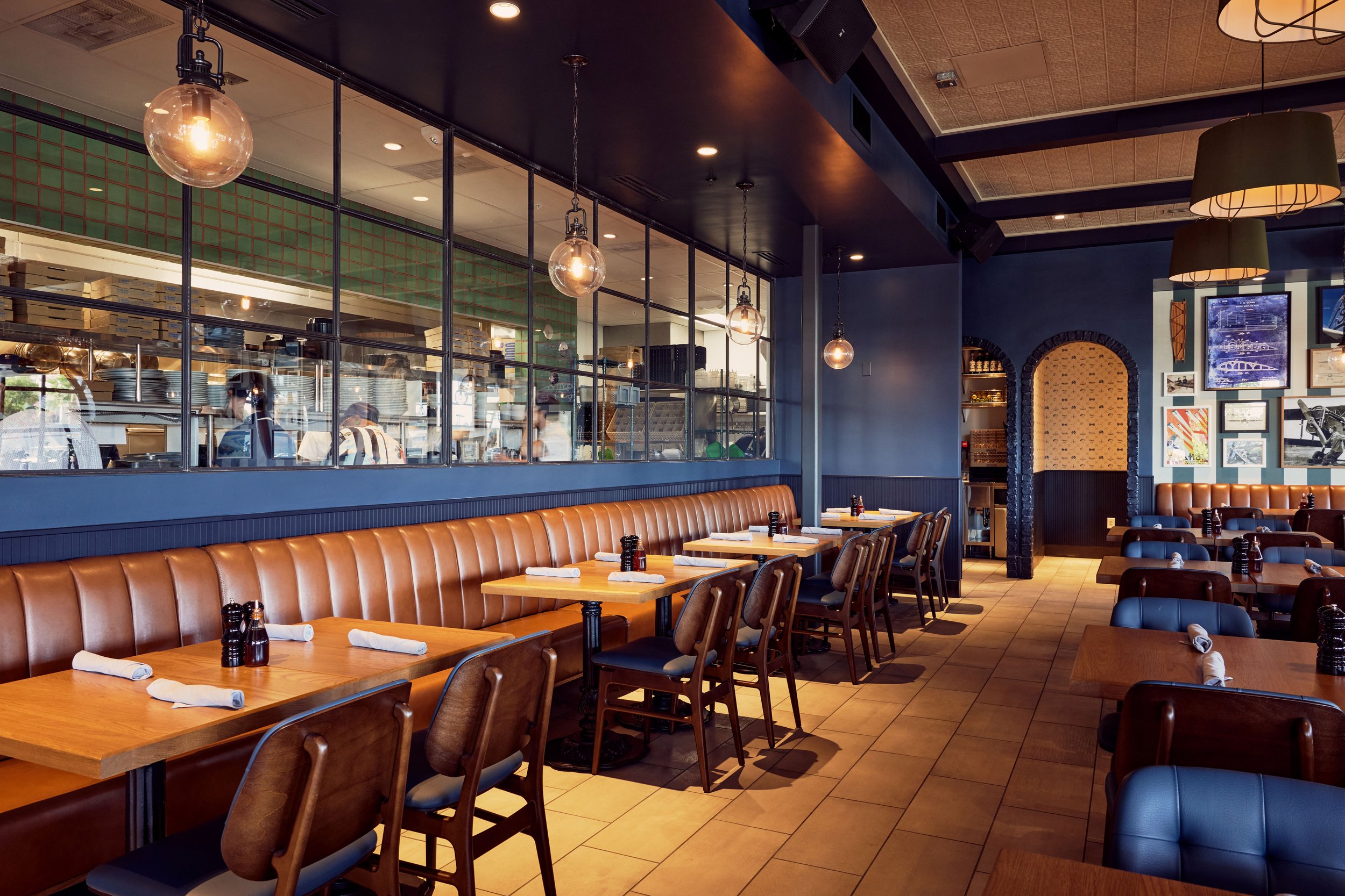 Empty dining area in a restaurant with wooden tables, leather chairs, and framed artwork on blue walls, with a view into the open kitchen through a glass partition.