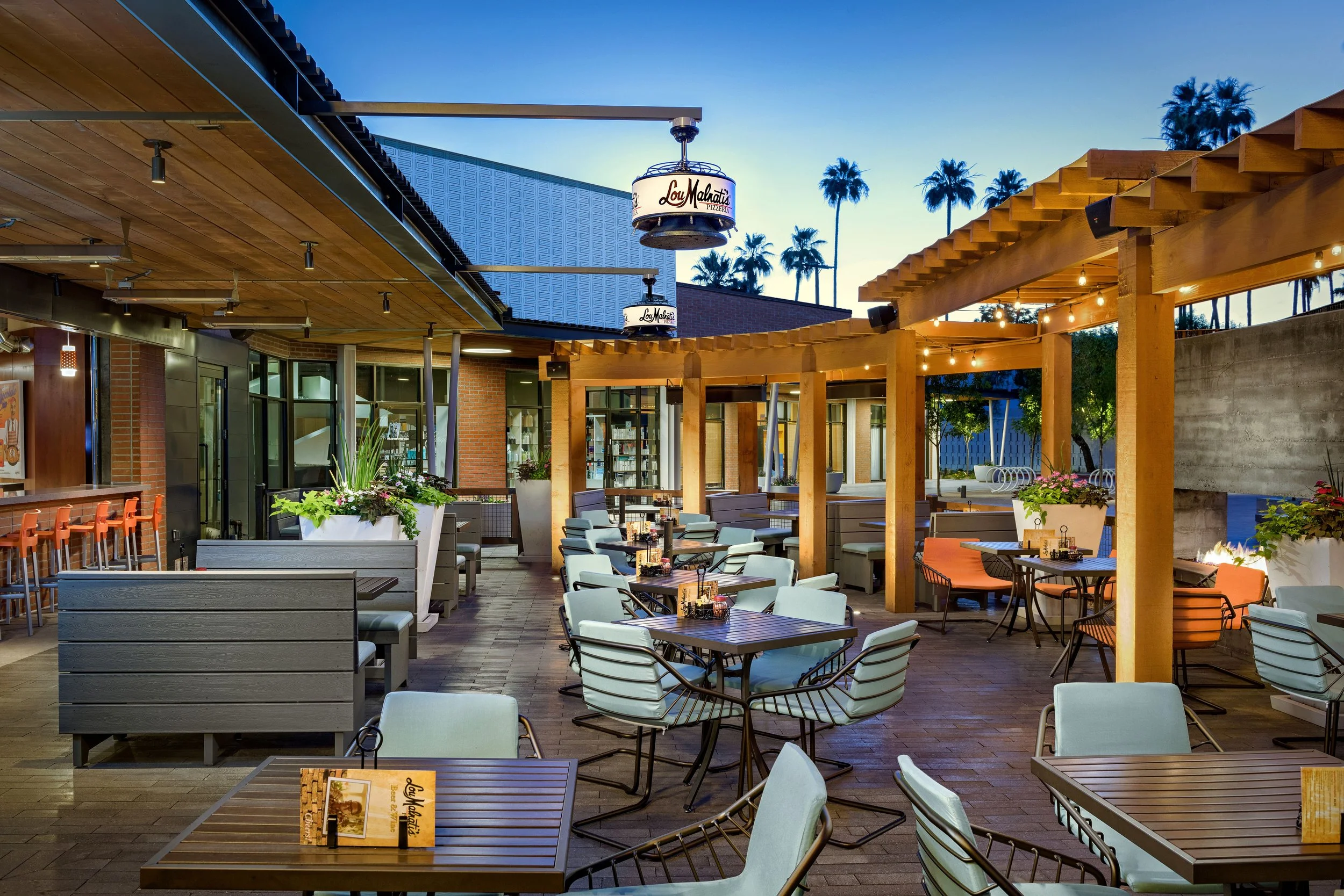 An outdoor dining patio with tables and chairs, decorative string lights, potted plants, and a cityscape background with palm trees at dusk.