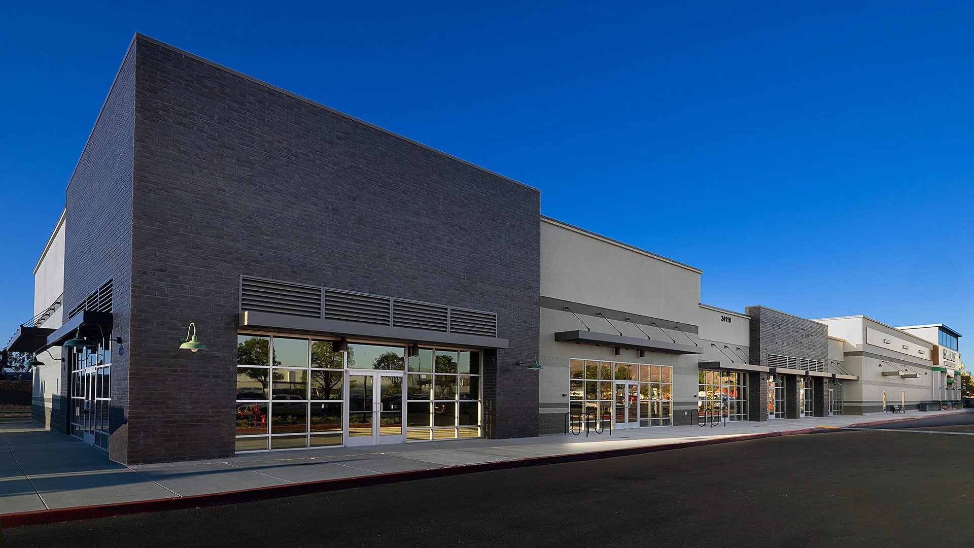 Exterior view of a modern shopping center with large glass storefronts, black brick walls, and awnings.