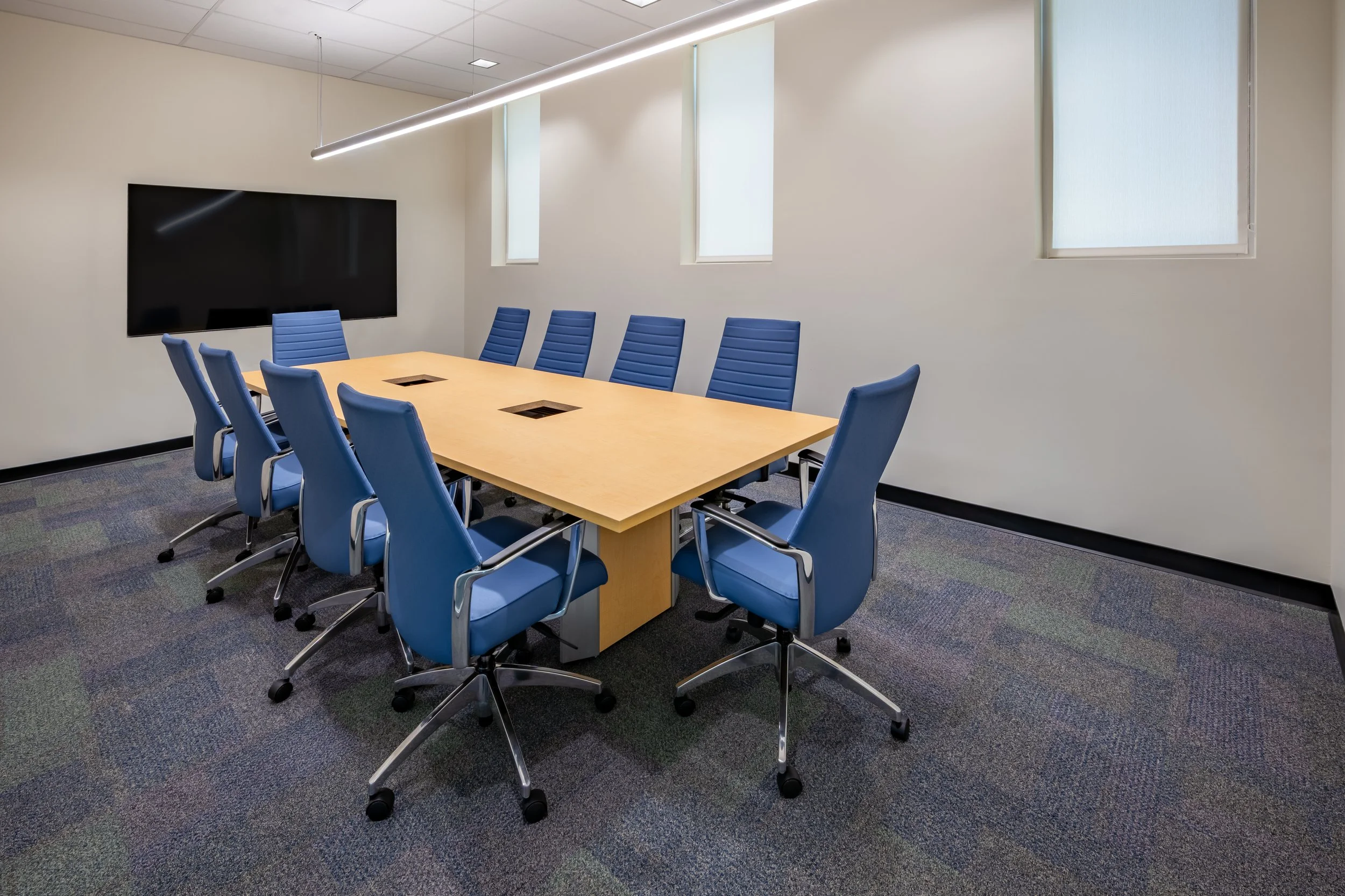 A modern conference room with a rectangular wooden table surrounded by ten blue office chairs with armrests and casters. The room has a large flat-screen television mounted on the wall and three windows with frosted glass letting in natural light. Th