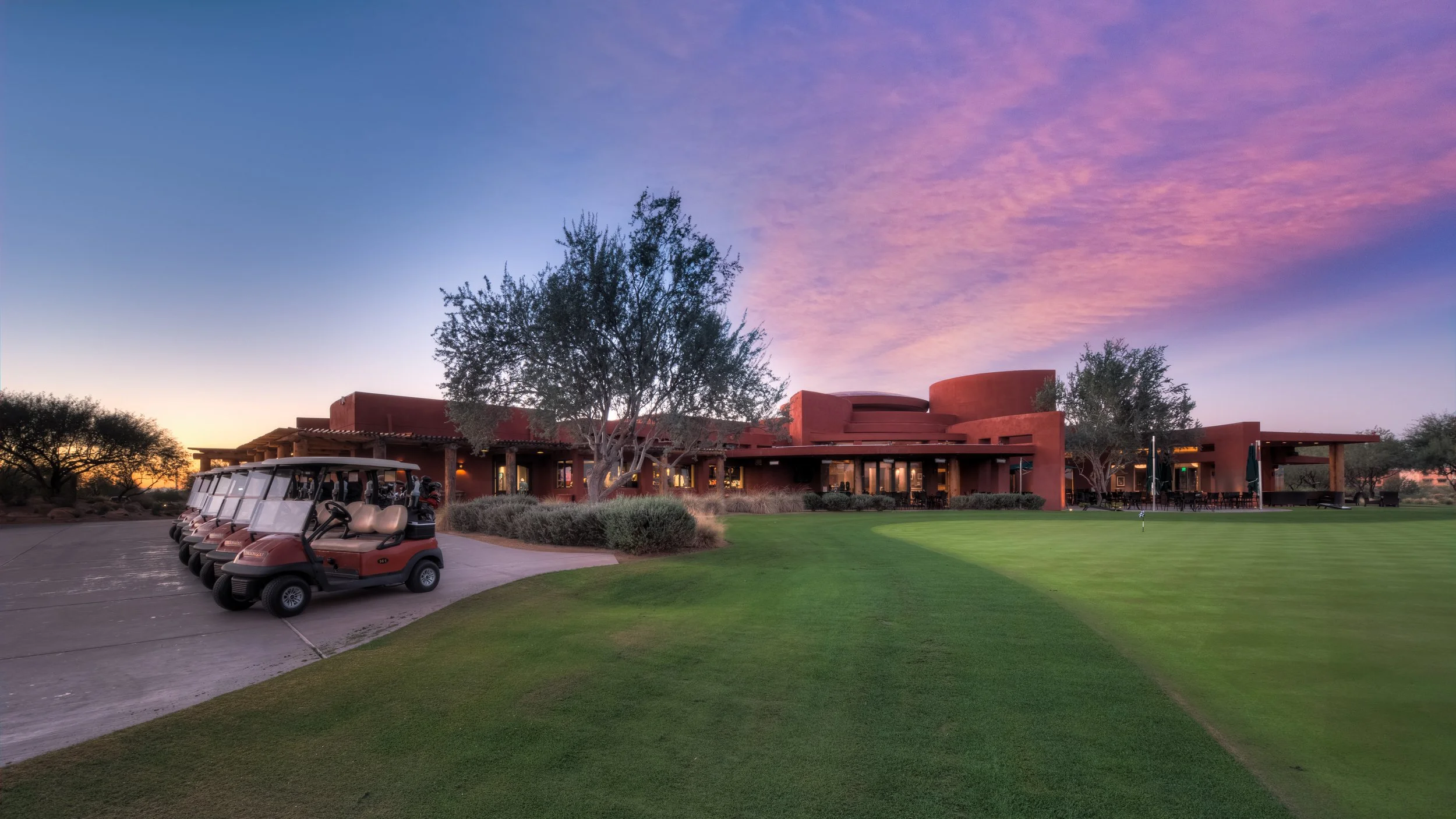 A golf course with neatly trimmed grass and a row of golf carts parked on a paved driveway. In the background, there is a reddish-brown building with outdoor seating under a covered patio. Trees surround the area, and the sky displays a vibrant pink 
