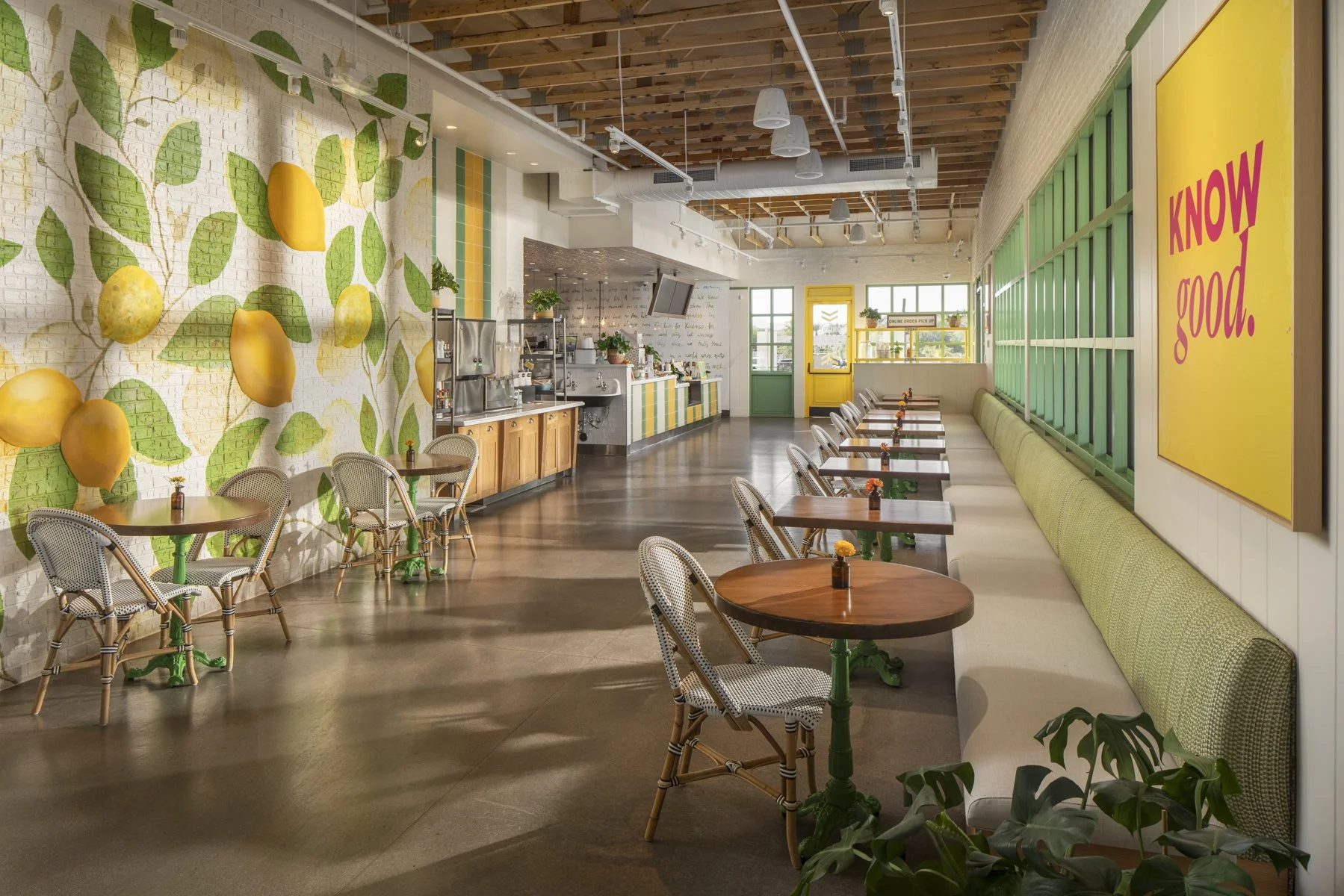 Bright cafe interior with a mural of a lemon tree on the wall, wooden tables with chairs, and a colorful counter area. Large windows, green accents, and a yellow sign that says 'KNOW good.'