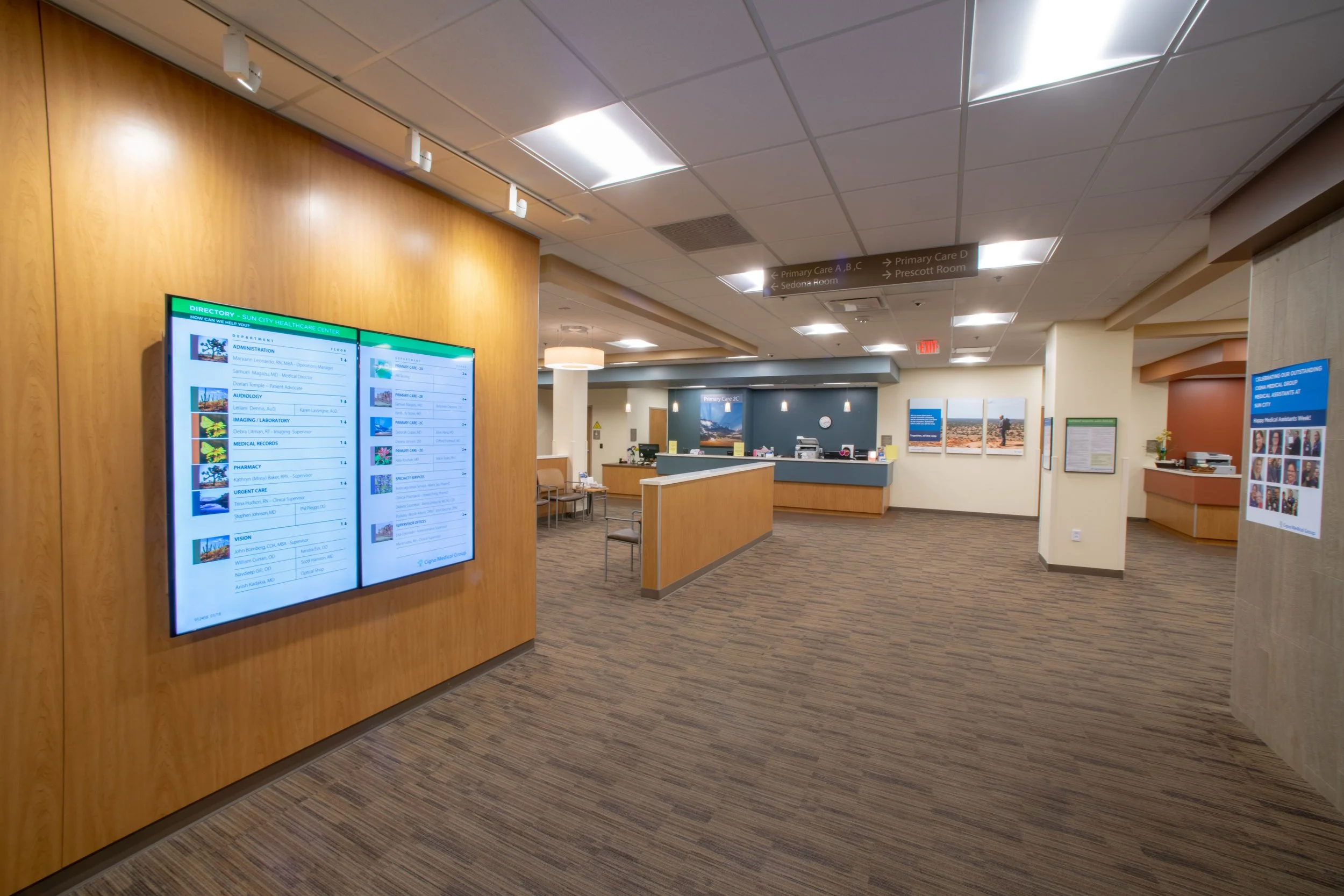 Hospital reception area with a digital directory screen on a wooden wall, a reception desk in the background, informational posters on the walls, and ceiling signs directing to primary care and other departments.