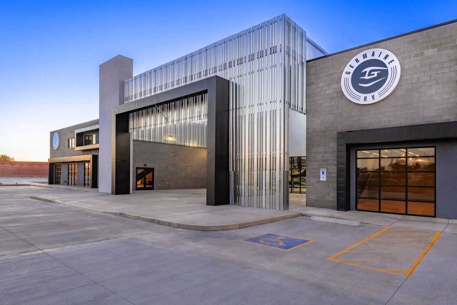 Modern building with a sign that reads 'Germain RV,' featuring a large entrance framed by vertical metallic slats and a handicapped parking space in the foreground during early evening.