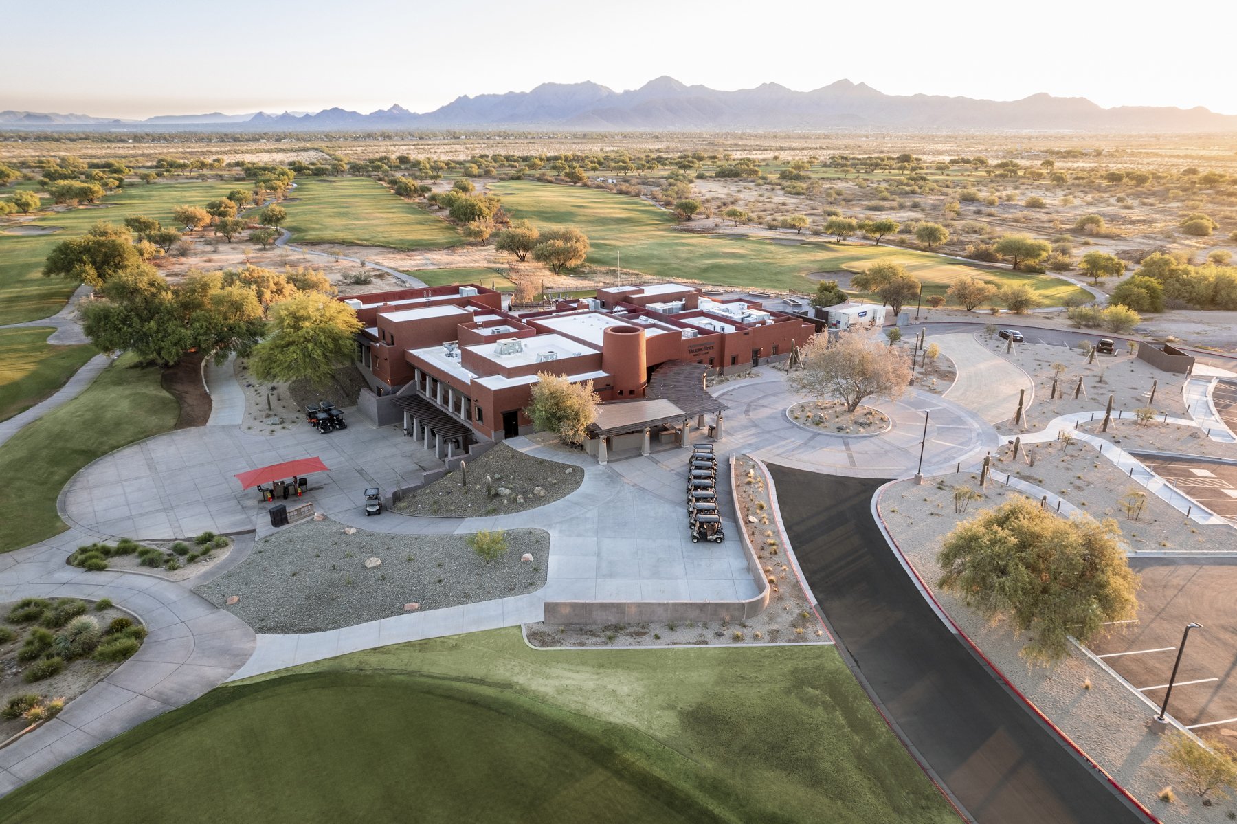 Aerial view of a desert golf course with a clubhouse, parking lot, and surrounding sand dunes and mountains in the background.