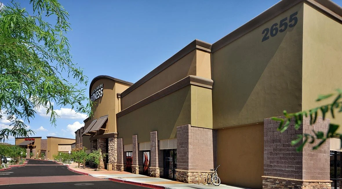 Empty shopping mall parking lot with trees, a bicycle, and storefronts with signs, in sunny weather.