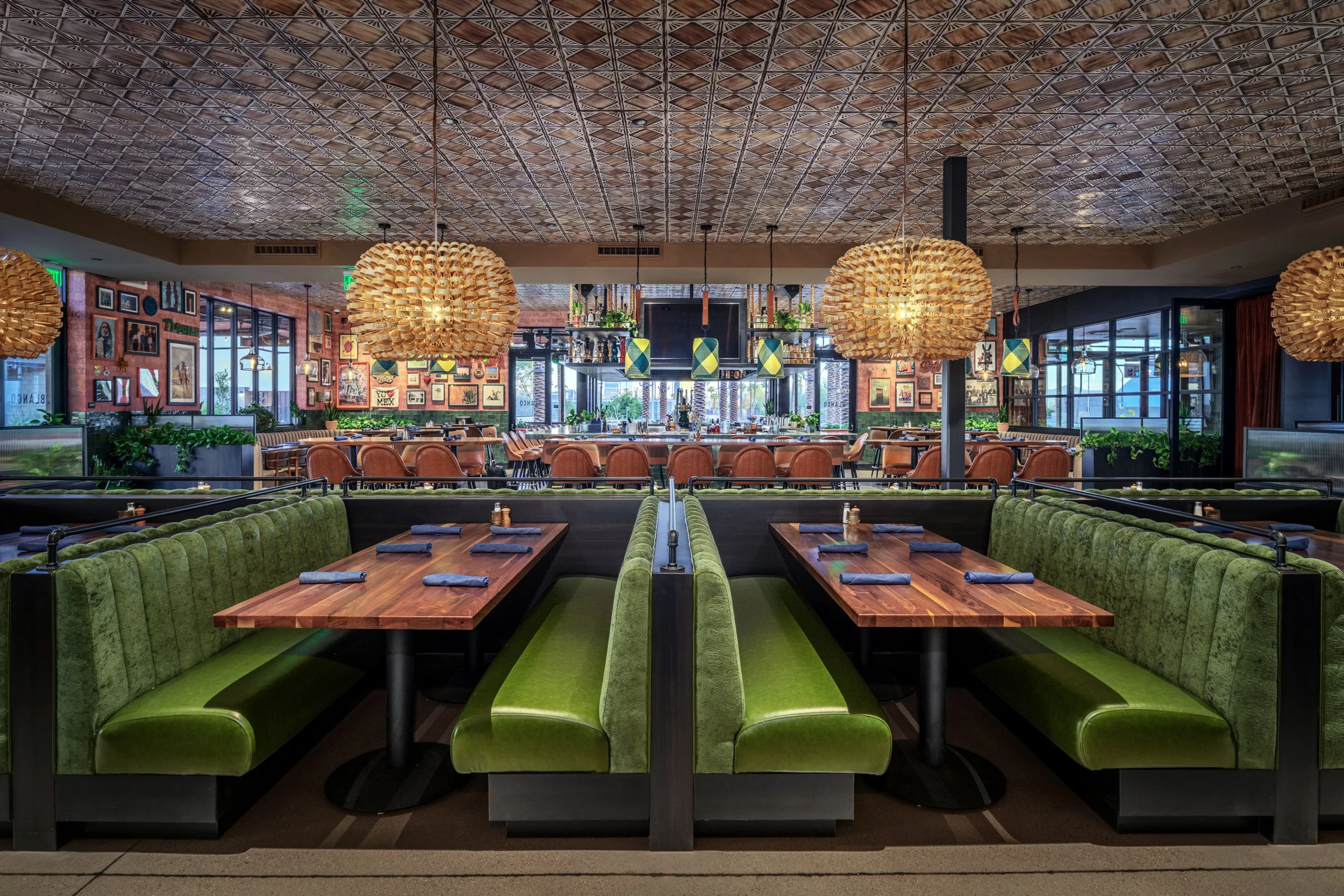 Interior of a restaurant with green booth seating, wooden tables, and hanging pendant lights, decorated with framed artwork on brick walls and large windows.