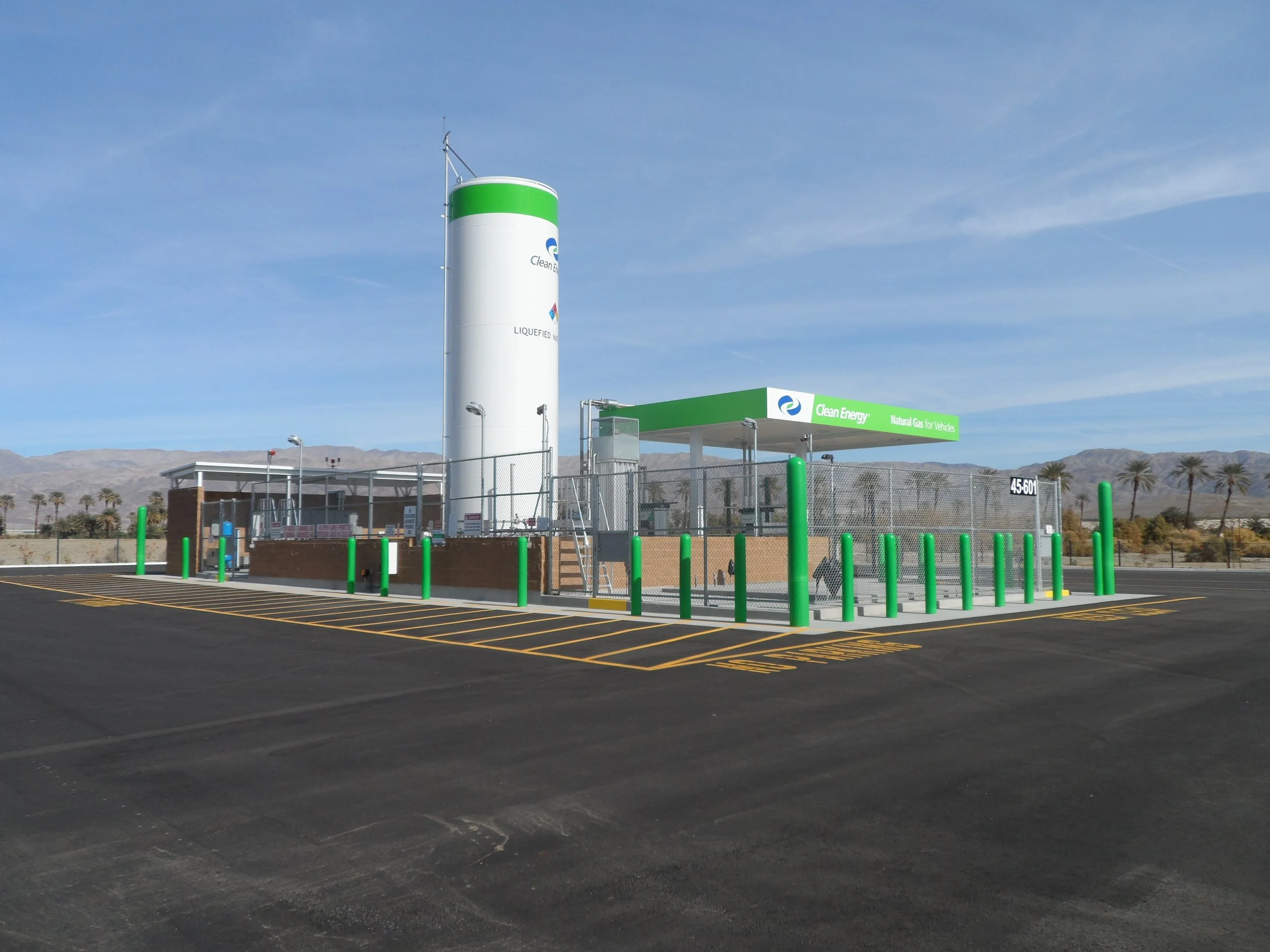 A clean energy refueling station for natural gas vehicles situated in a desert landscape with mountains and palm trees in the background, featuring green and white signage and safety bollards.