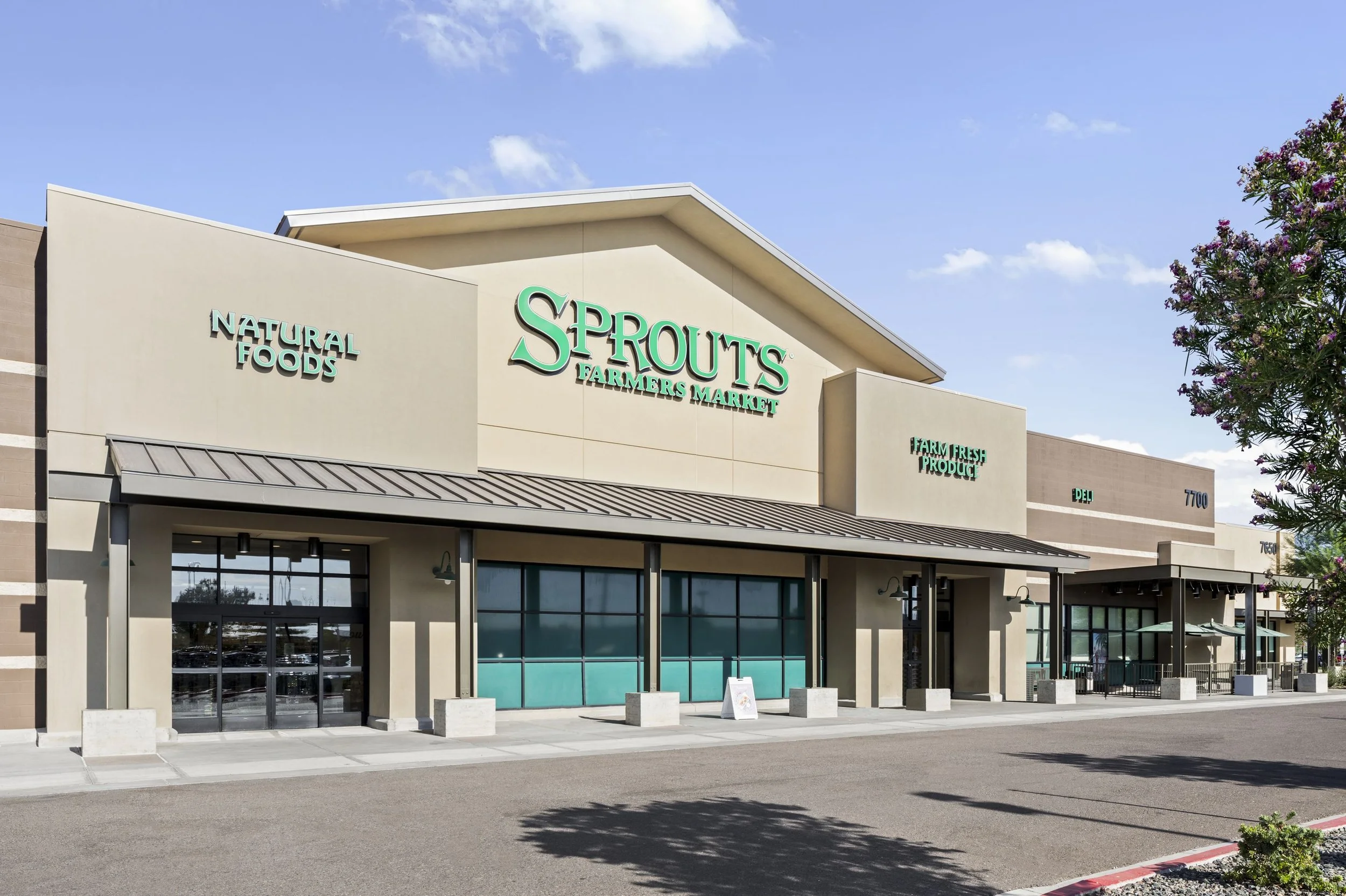 Exterior view of Sprouts Farmers Market store with signage for natural foods, farm fresh products, and deli, under a blue sky with few clouds and pink flowering tree on the right.