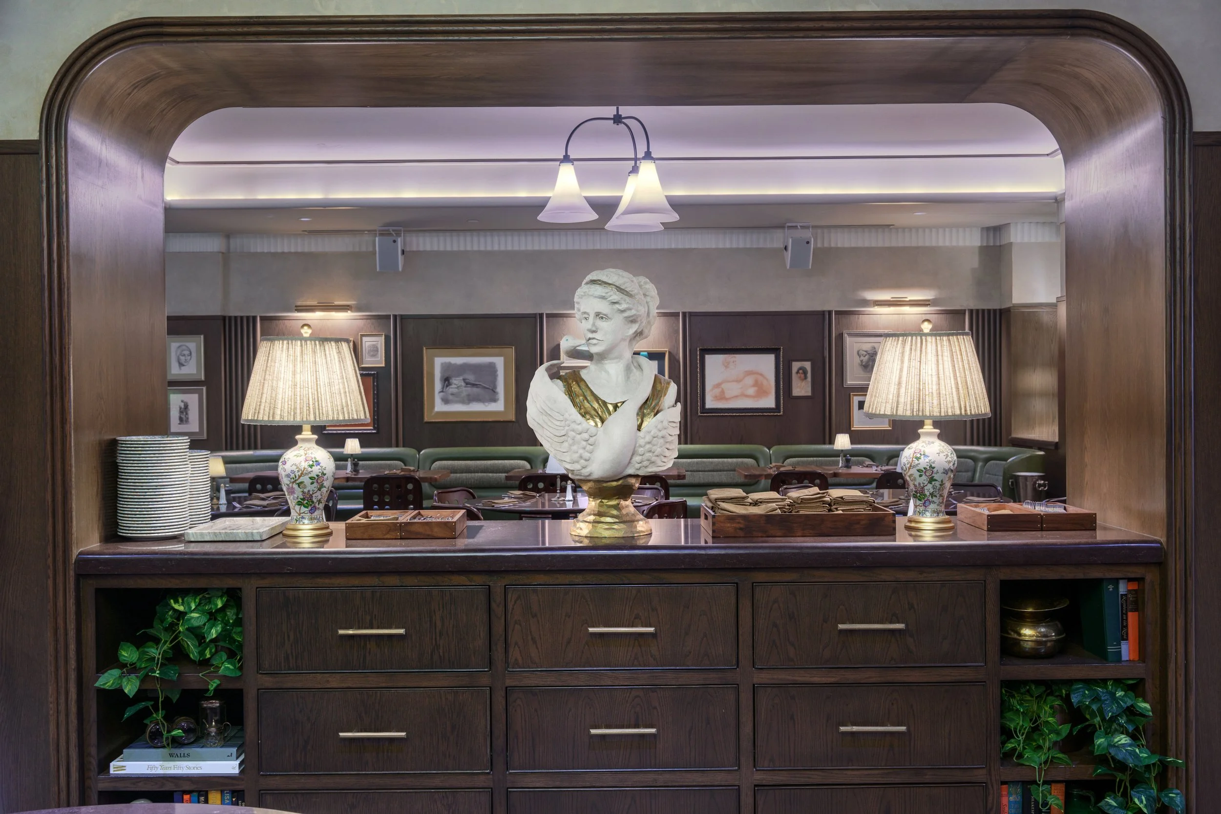 View of a restaurant or dining area with a wooden counter in the foreground, featuring a bust sculpture of a woman on a pedestal, flanked by two lamps. Behind the counter, there are tables with plates and napkins, framed artwork on the walls, and row