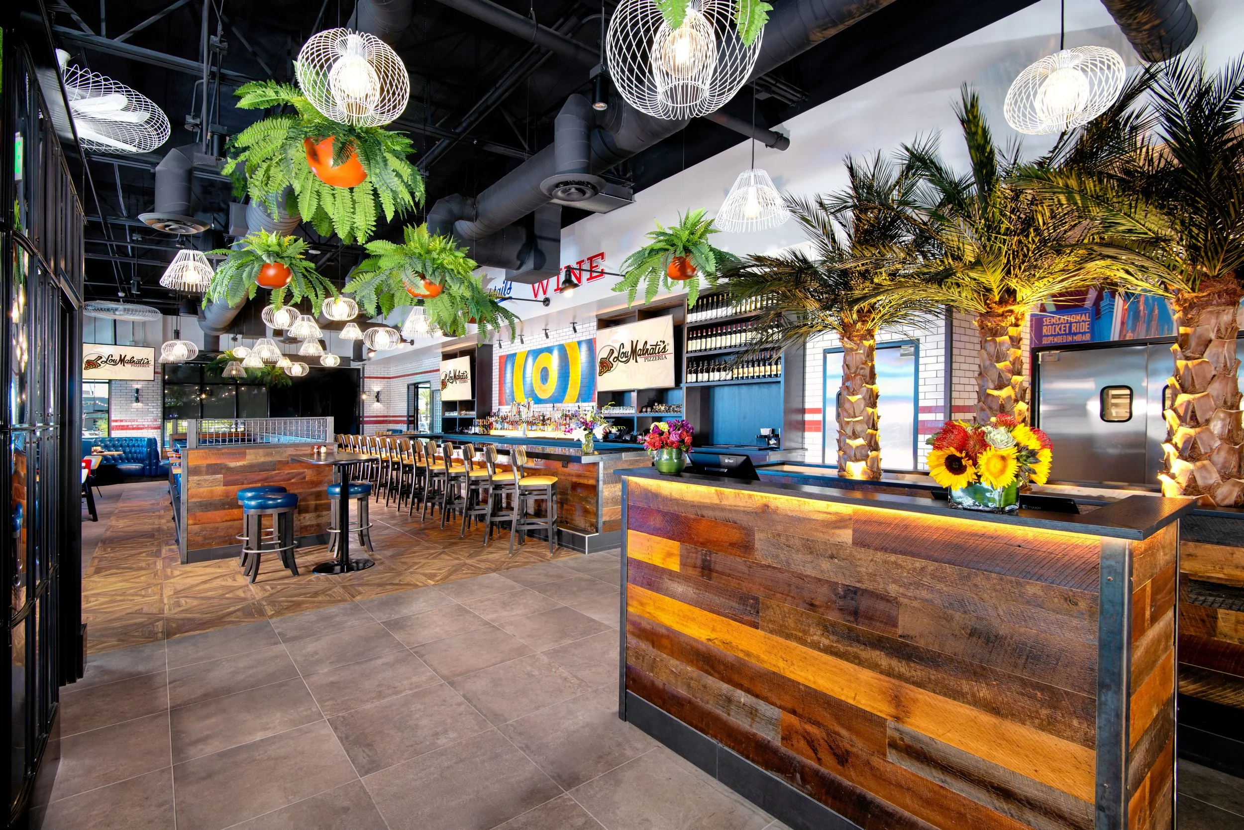 Interior of a restaurant with tropical decor, including hanging fern plants with orange pots, palm trees, and bright lighting fixtures. There is a wooden bar with flowers and a seating area with chairs and tables.