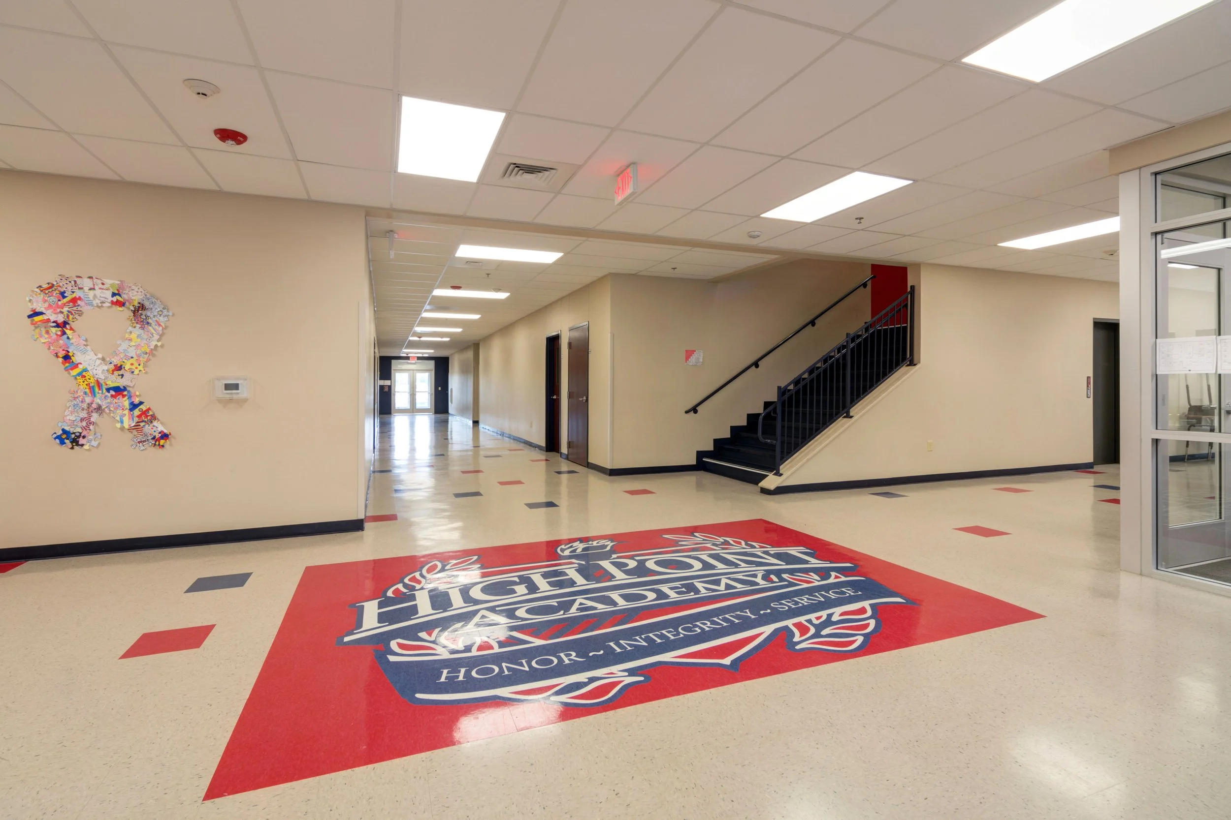 School hallway with a large painted logo on the floor that reads 'High Point Academy' with the words 'Honor - Integrity - Service' underneath. The hallway features beige walls, black baseboards, various doors, and a staircase. There is a colorful pin