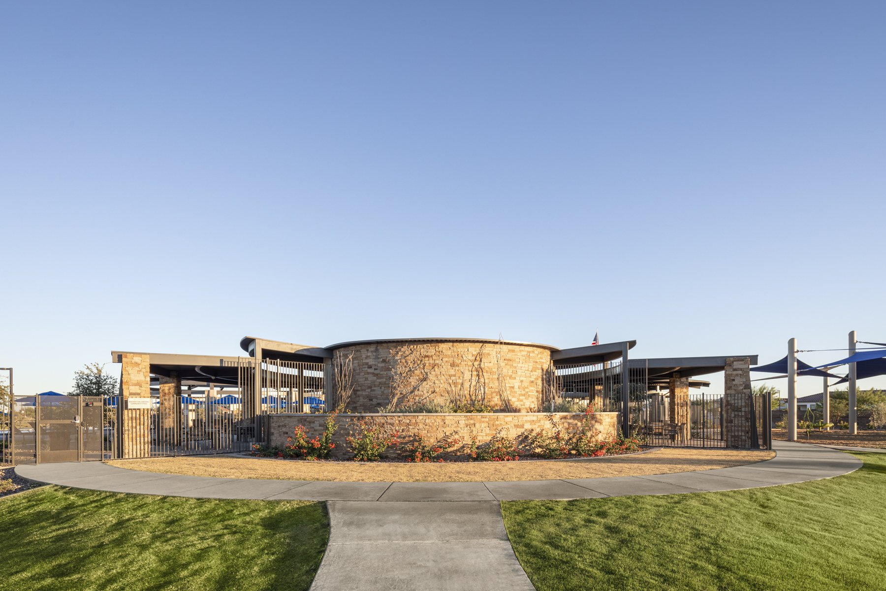 A modern park pavilion with brick walls, metal fences, and blue shade structures, surrounded by green grass and flower beds, under a clear blue sky.