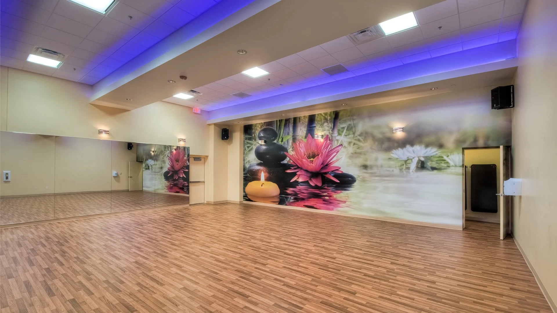 Empty dance or yoga studio with hardwood floors, mirror wall, and a large mural of water lilies, stones, and candles on the back wall, blue ambient lighting on the ceiling.