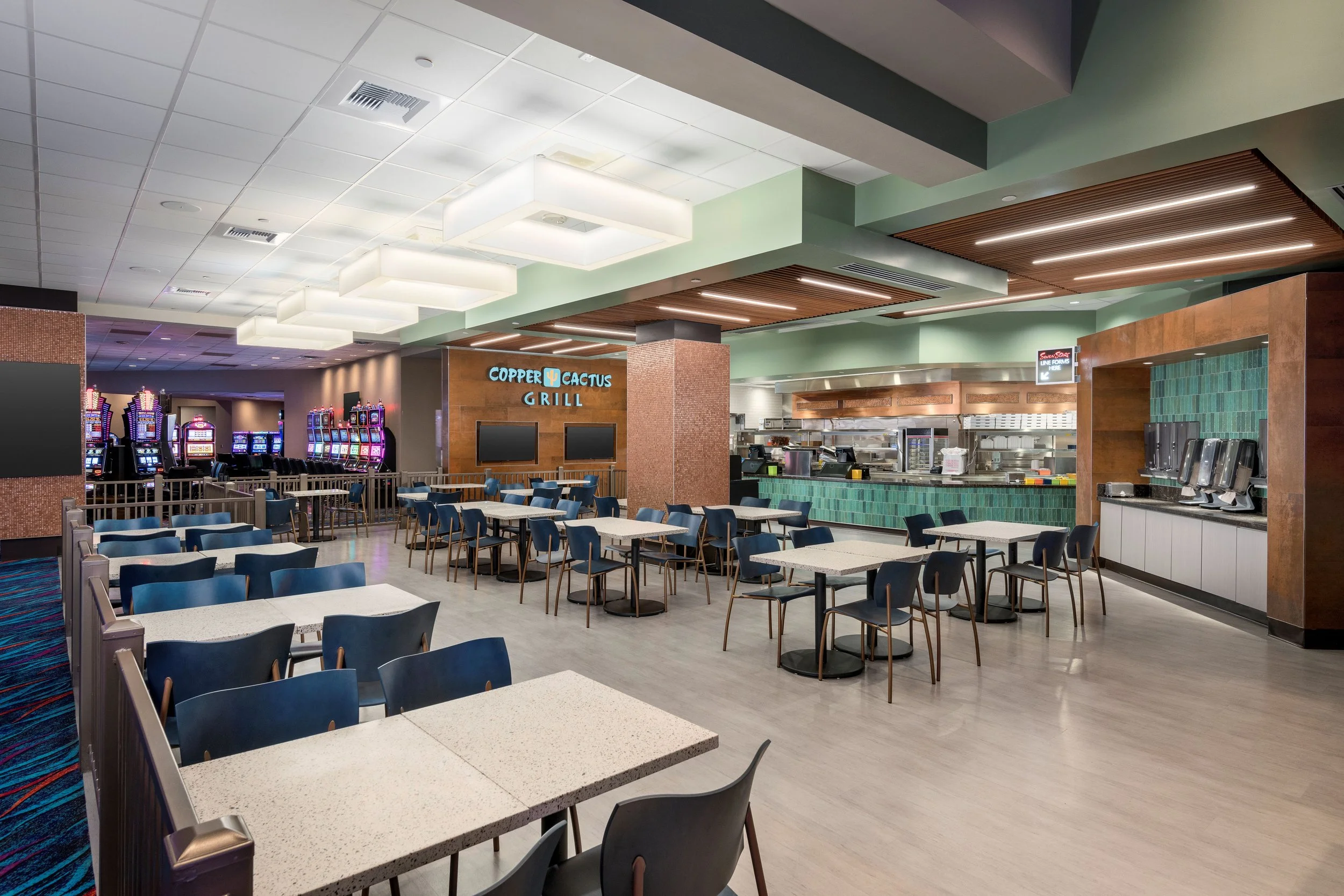 Empty restaurant with blue chairs and white tables, with gaming machines in the background, and a food counter labeled 'Copper Cactus Grill'.