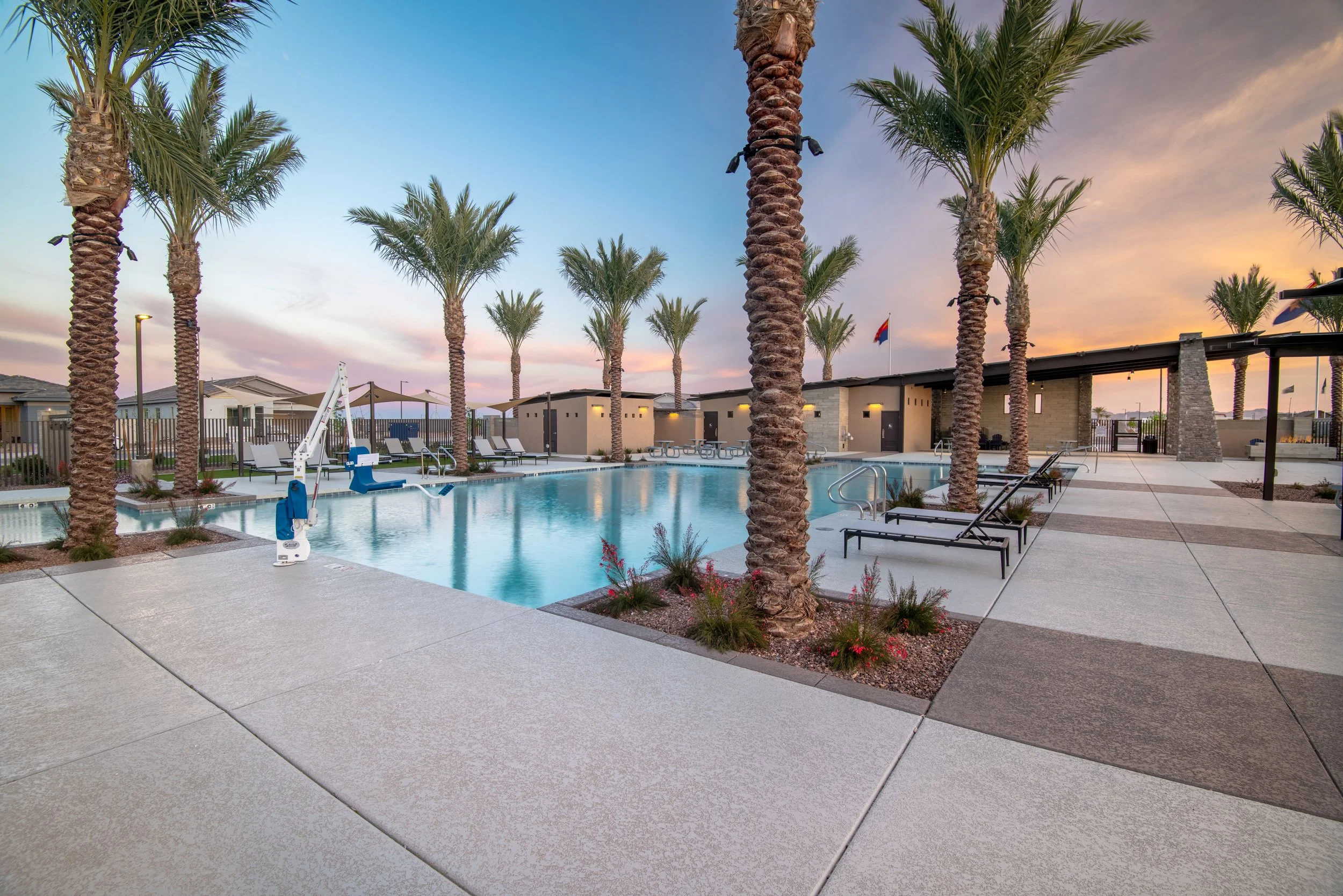 A swimming pool area with lounge chairs, palm trees, and nearby small structures at sunset.