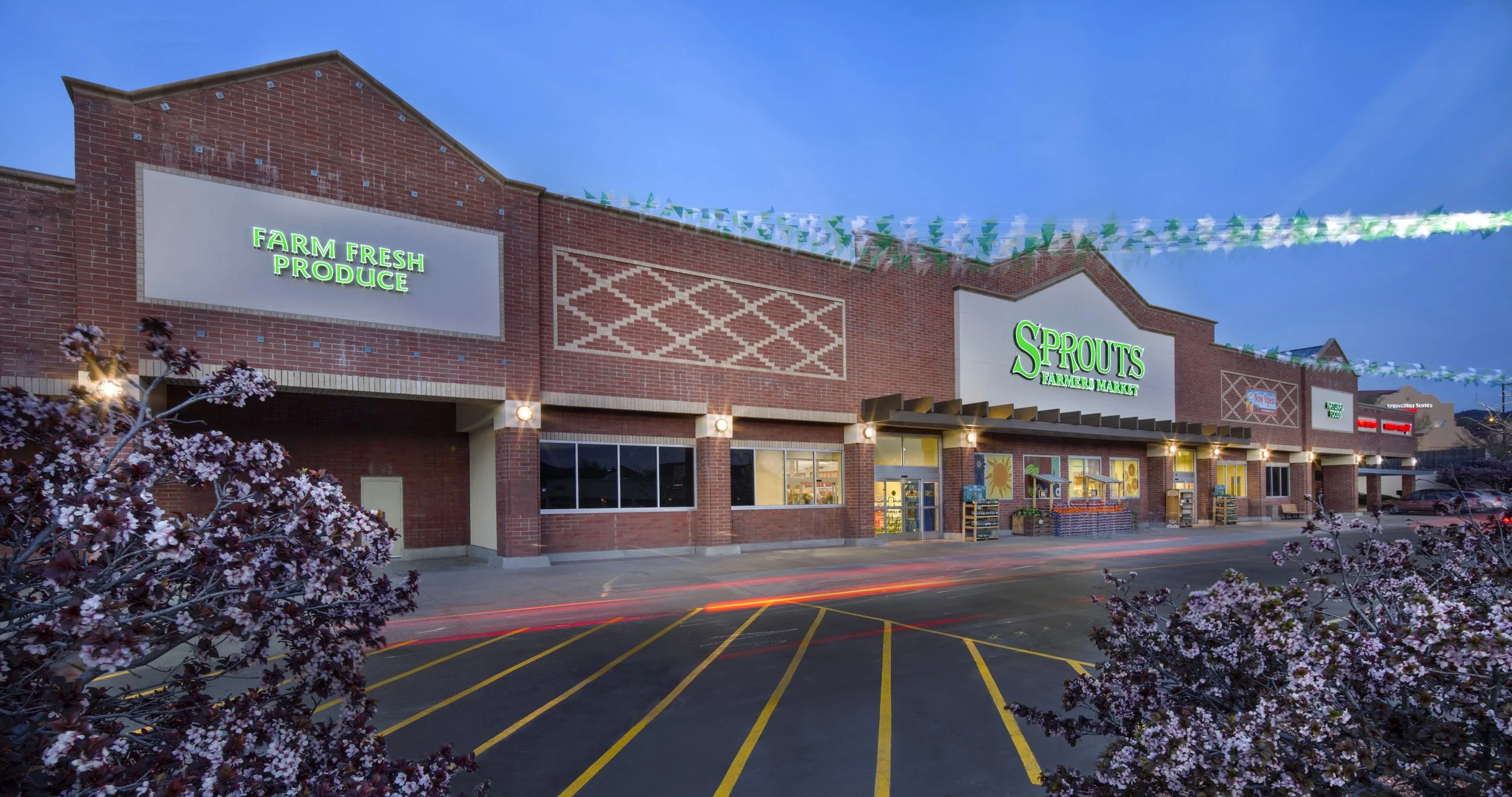 Exterior of a grocery store with signs for 'Farm Fresh Produce' and 'Sprouts Farmers Market' at dusk, with floral bushes in the foreground and a parking lot.
