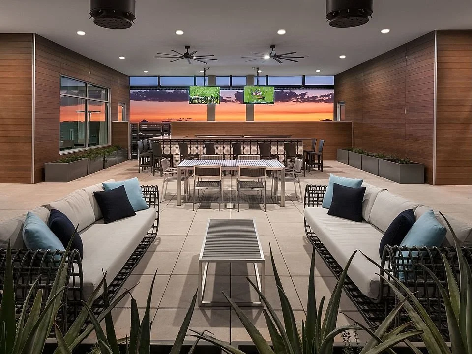 A spacious indoor lounge with large windows showing a sunset sky, modern furniture including sofas with cushions, a long dining table with chairs, and potted plants along the walls.