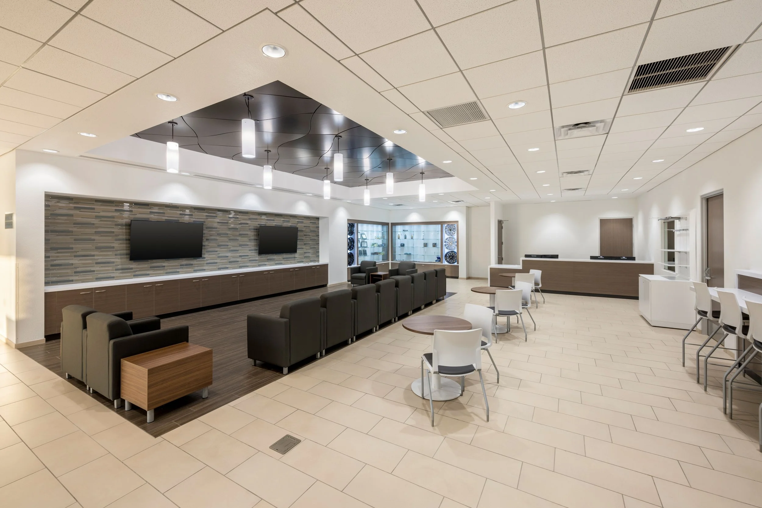 Modern waiting area with chairs, tables, a reception counter, and two large flat-screen TVs on a tiled wall.