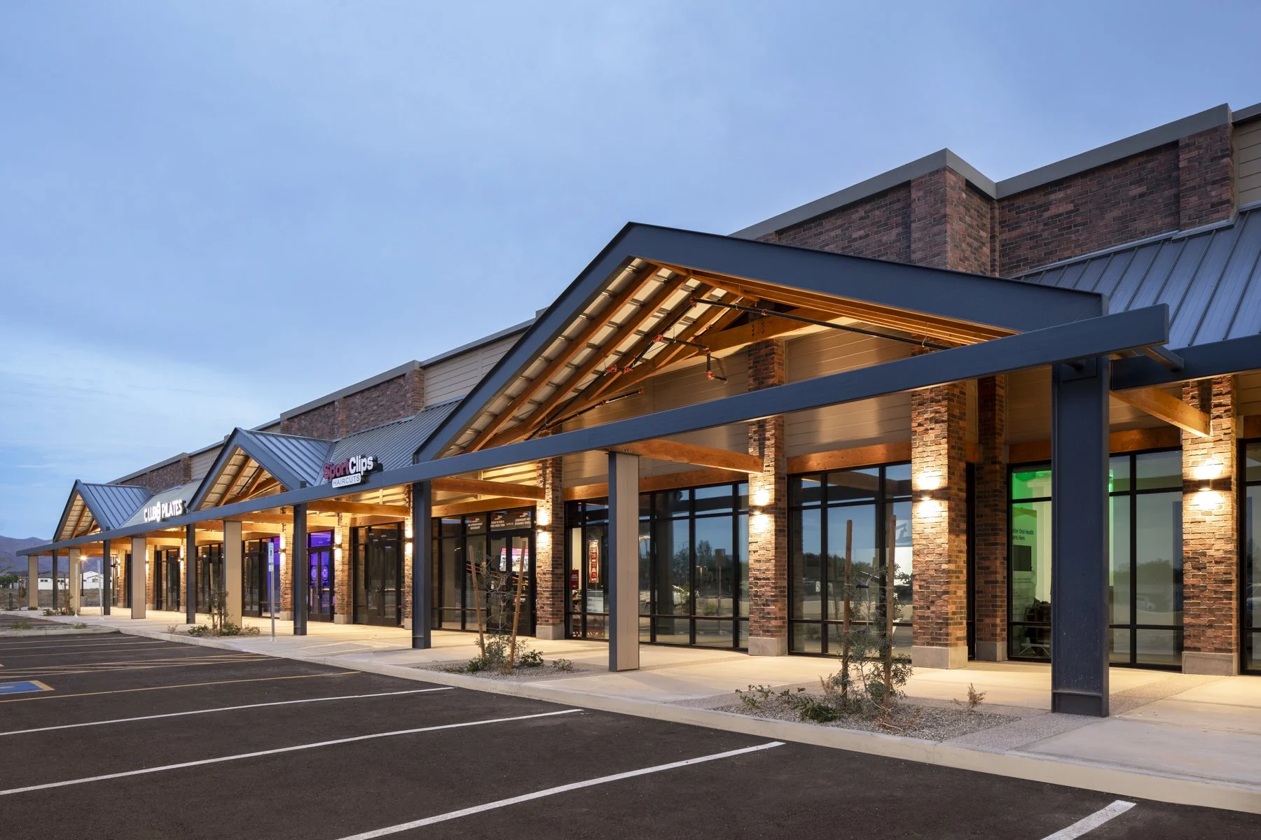 Exterior of a modern retail strip mall with parking spaces in the foreground, illuminated by exterior lights at dusk.