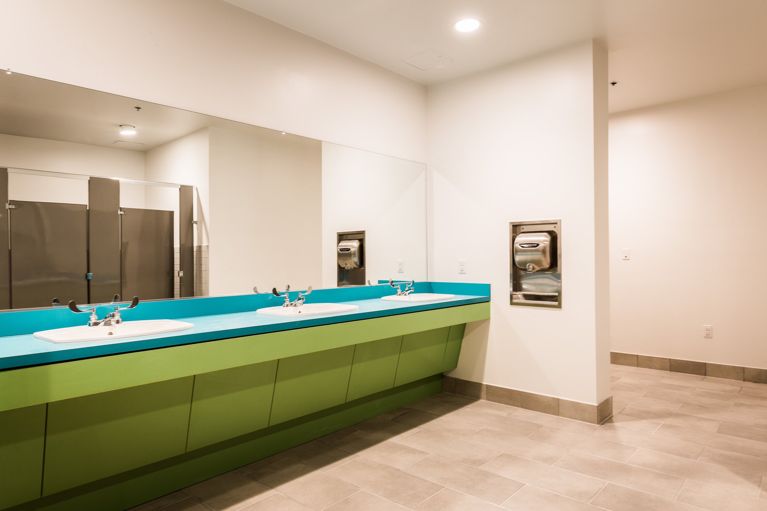 Public restrooms with three sinks on a blue counter, a large mirror, and stainless steel hand dryers on white walls, with beige tile flooring.