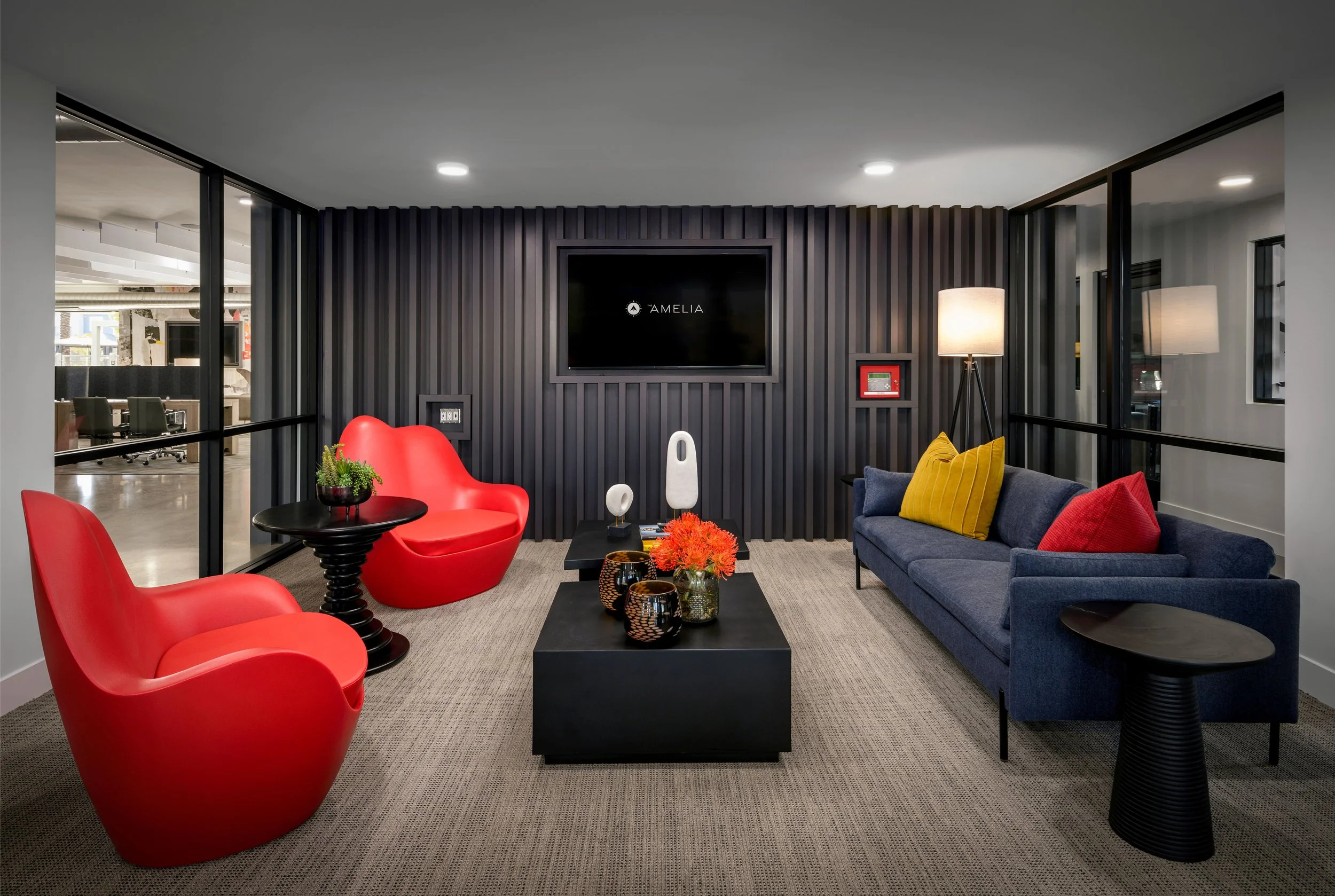 Modern office lounge with black textured wall, large TV screen displaying 'Amelia', colorful chairs, sofa with cushions, coffee table with decorative items, floor lamp, and glass walls showing nearby workspace.