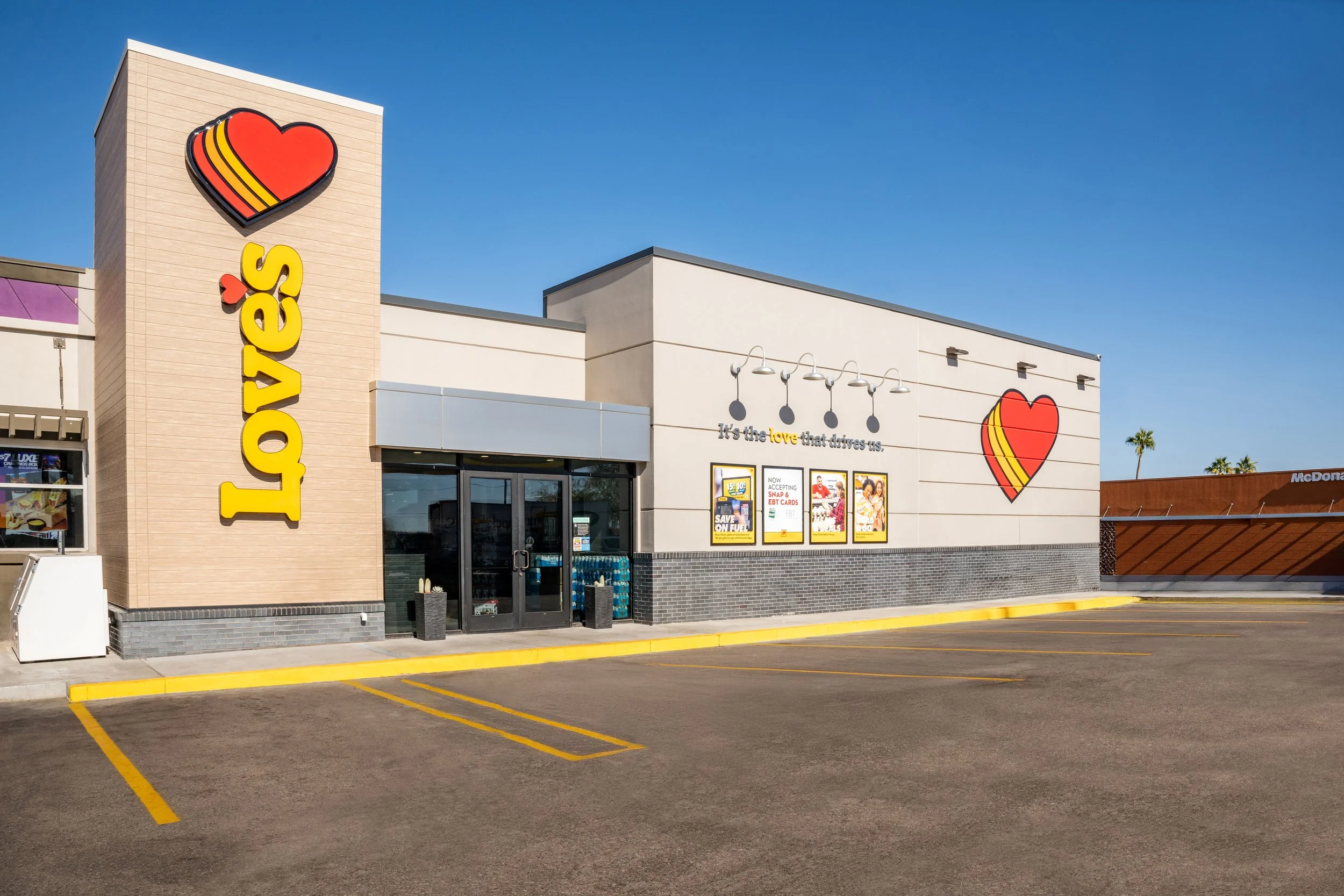 Exterior of a Taco Bell restaurant with a beige and white facade, a large yellow 'Taco Bell's' sign, and heart-shaped logo designs on the walls. Empty parking spaces in front, clear blue sky, and a McDonald's sign visible in the background.