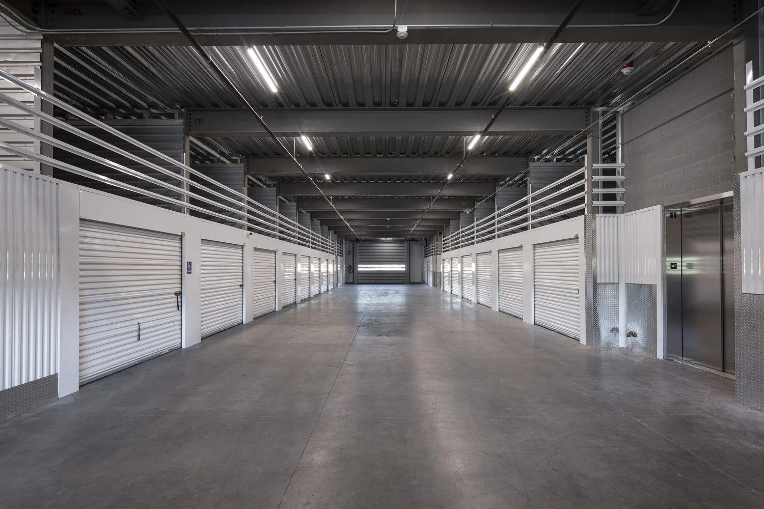 Empty indoor storage unit corridor with white roll-up doors on both sides, metal railings, polished concrete floor, and a partially open roll-up door at the far end.