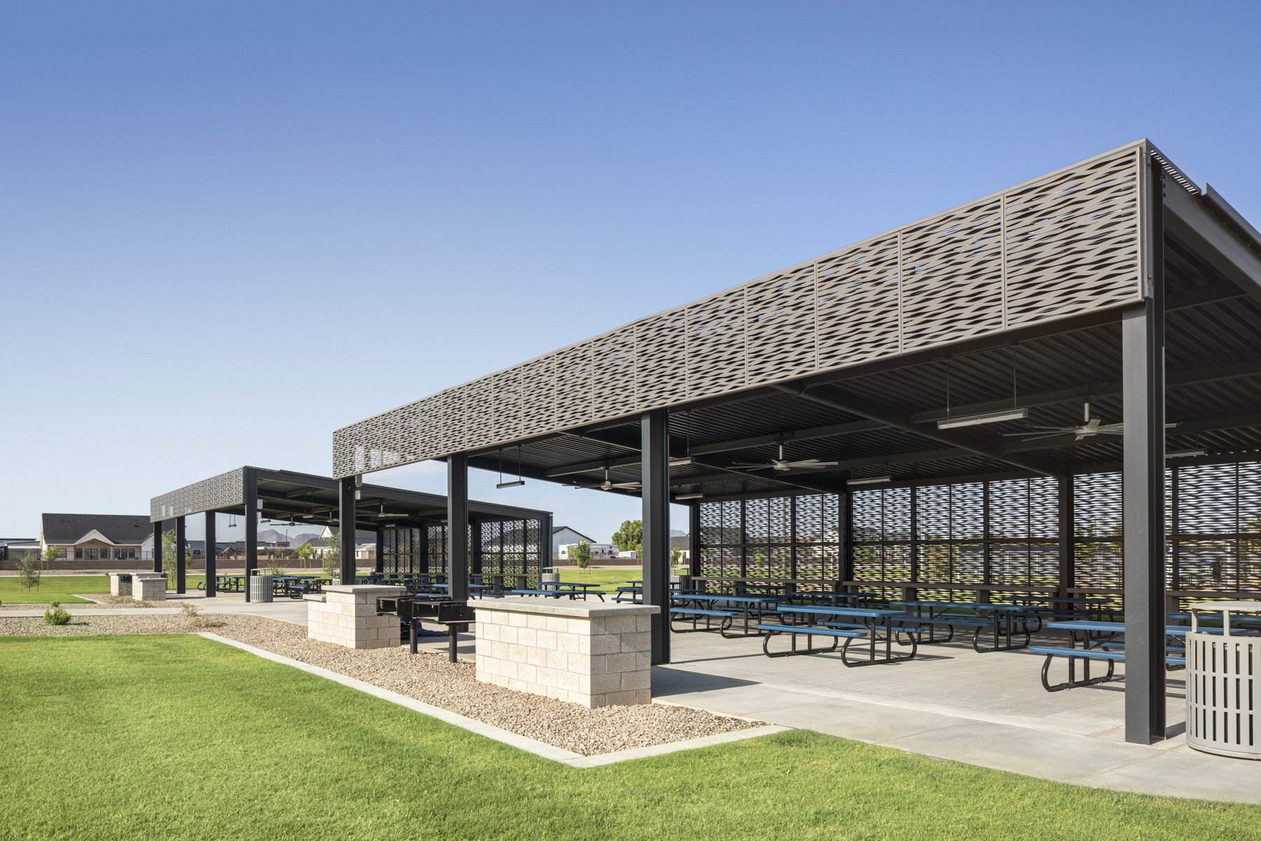 An outdoor picnic shelter with multiple picnic tables, surrounded by green grass and a park setting, under a clear blue sky.