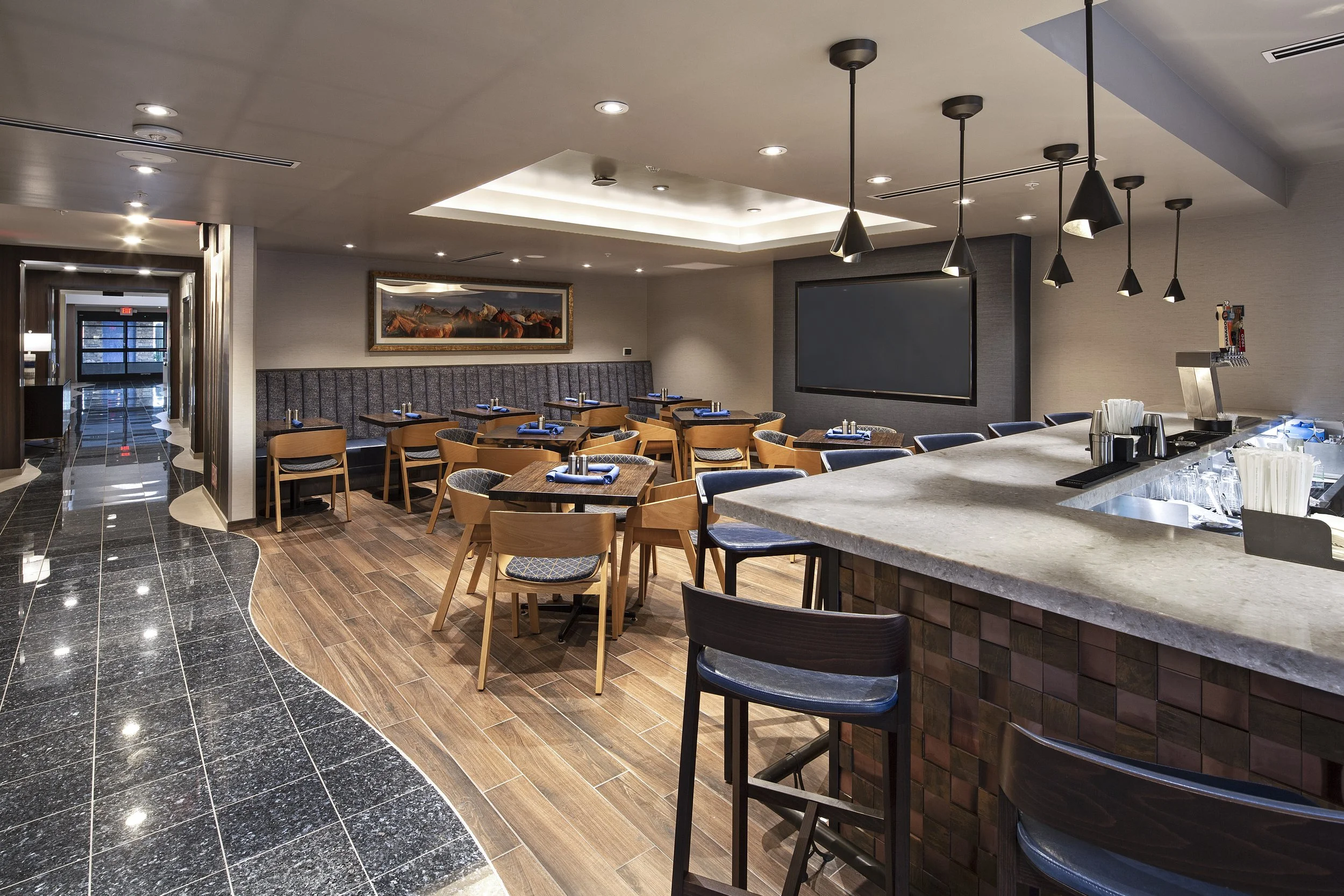A modern restaurant interior featuring a curved black tiled floor, a bar with stools, wooden dining tables with chairs, a large wall-mounted television, and a framed landscape painting on the wall.