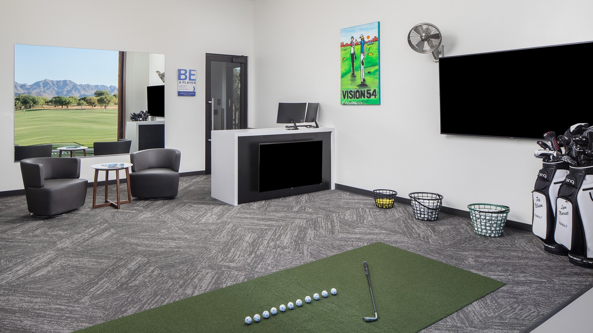 Golf simulator room with a green practice mat, a golf club, and a row of golf balls. Inside seating area with two chairs, a small table, and a view of a golf course through a window. Wall decorations include a colorful poster and a mirror. Golf bags 
