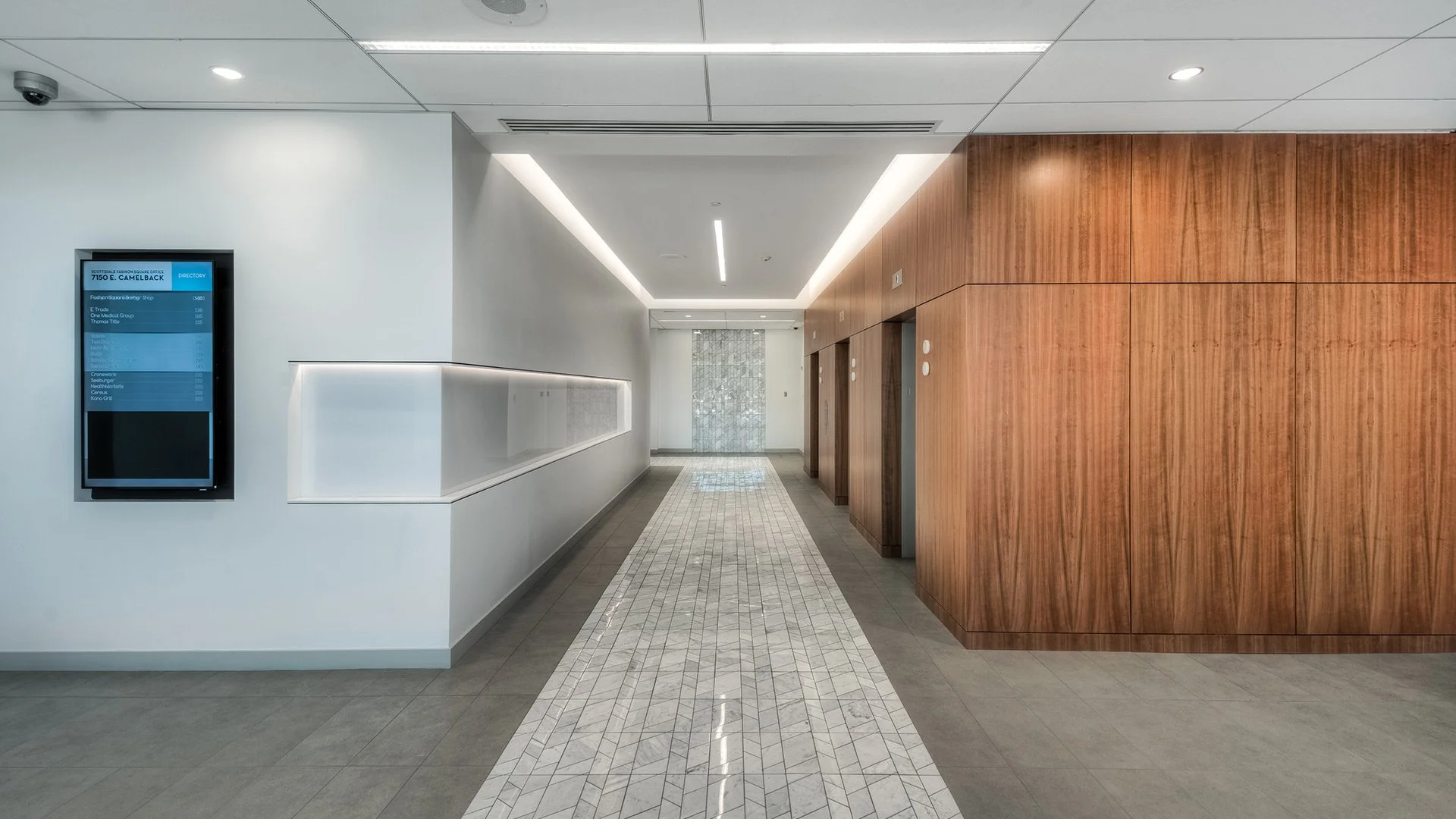 Modern indoor hallway with white and wooden walls, elevator doors, and a directional directory display on the left.