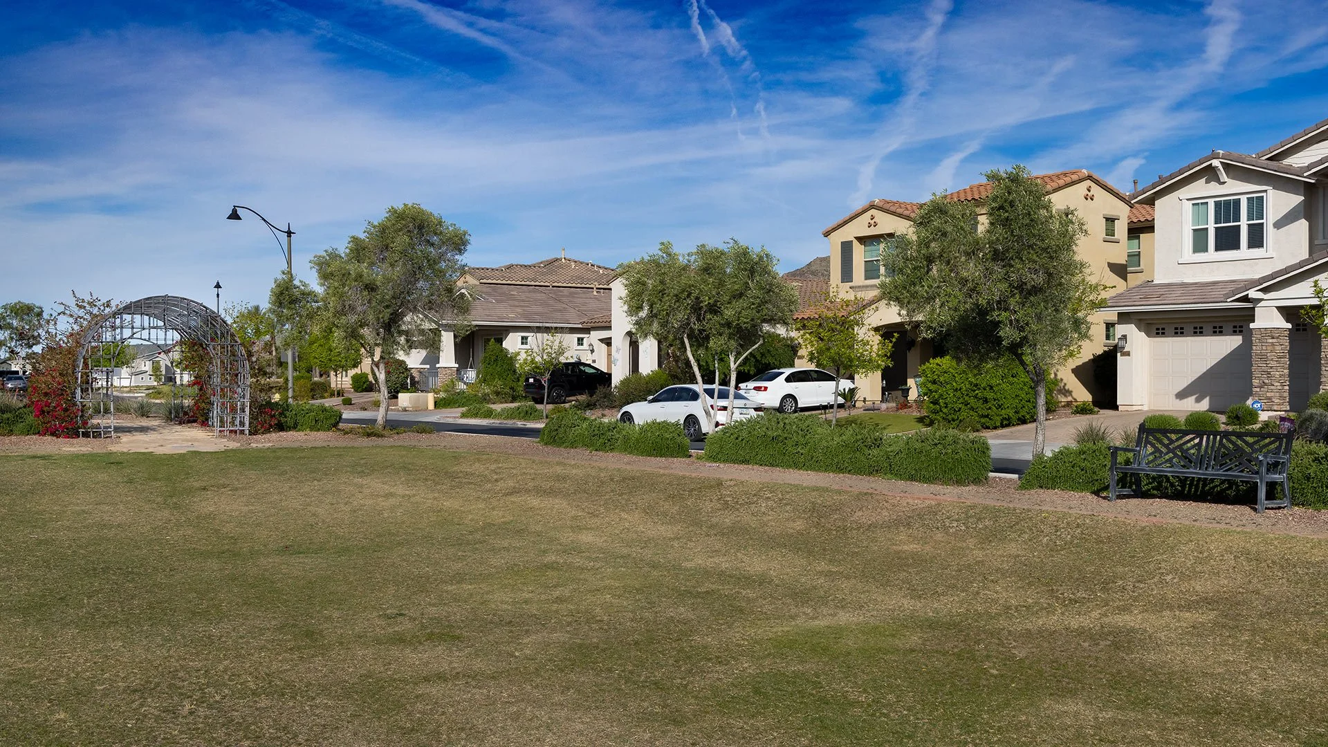 Suburban neighborhood with a grassy park, trees, houses, cars parked along the street, a black bench, and a decorative garden arch in the park under a blue sky with wispy clouds.