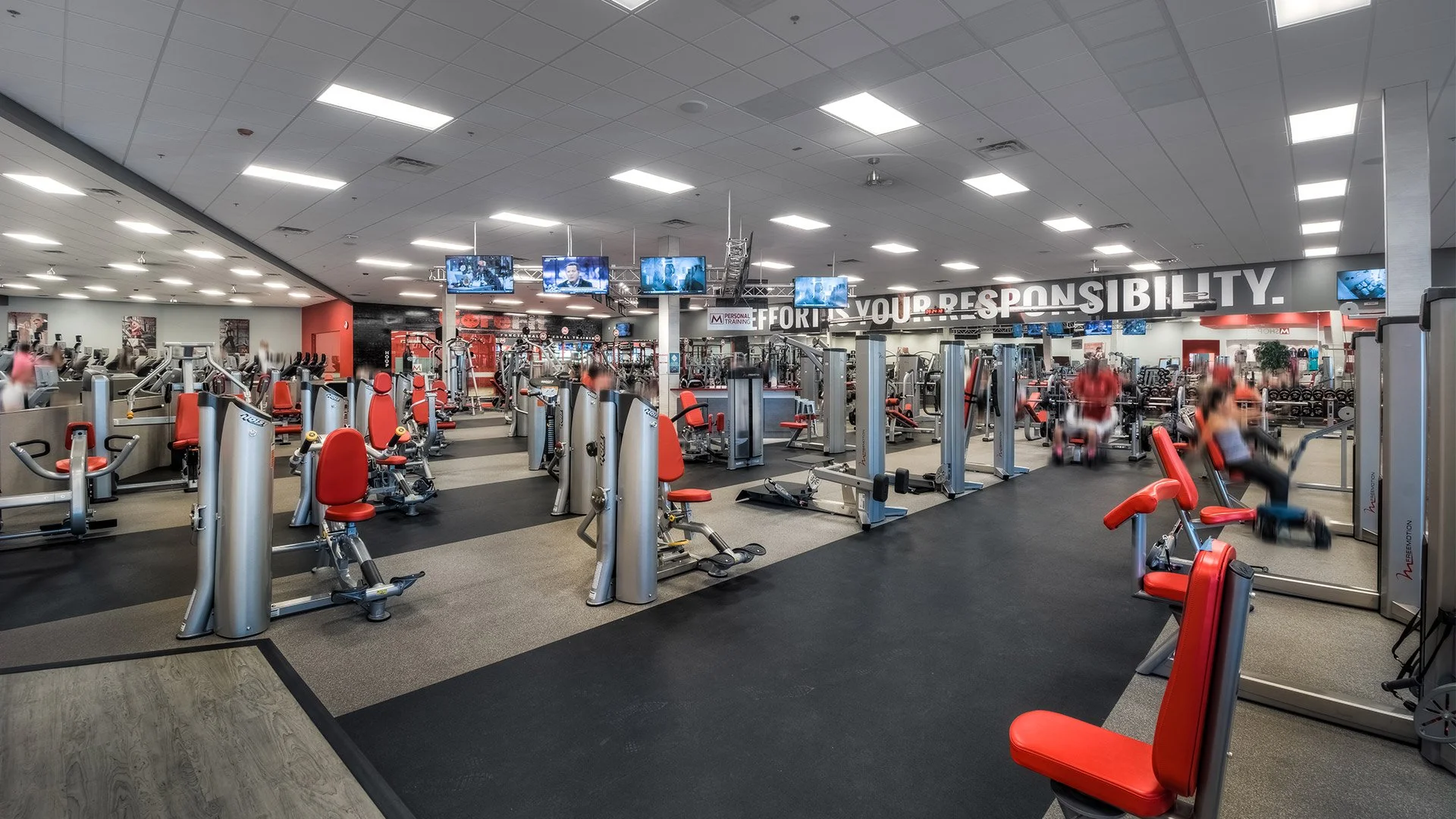Empty gym with various workout machines and equipment, with some people using the equipment, digital screens on the ceiling, and a large motivational quote on the wall.