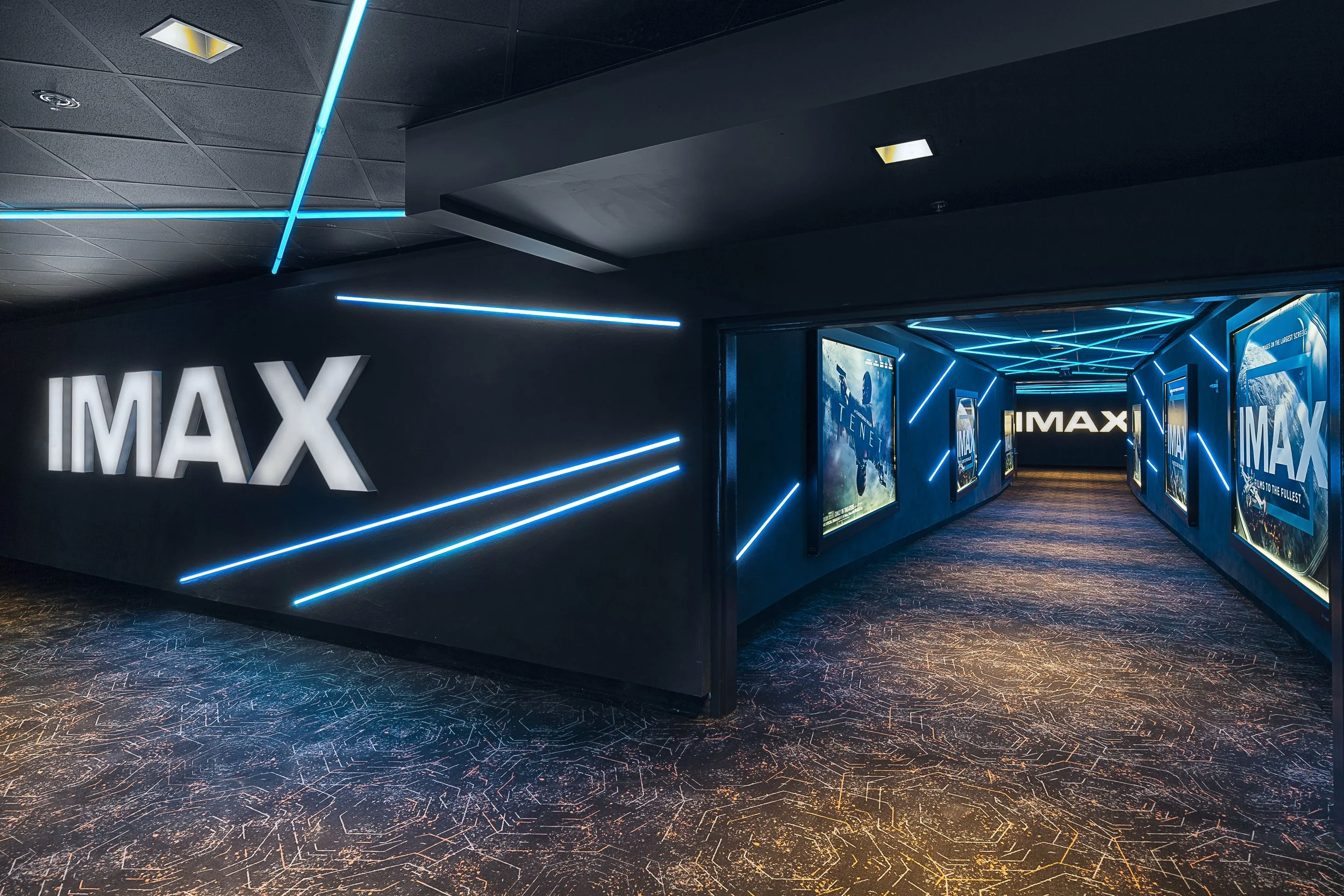 Interior of a modern movie theater with black walls, bright blue neon ceiling lights, illuminated 'IMAX' signs, and movie posters on the walls, leading into an IMAX screening area.