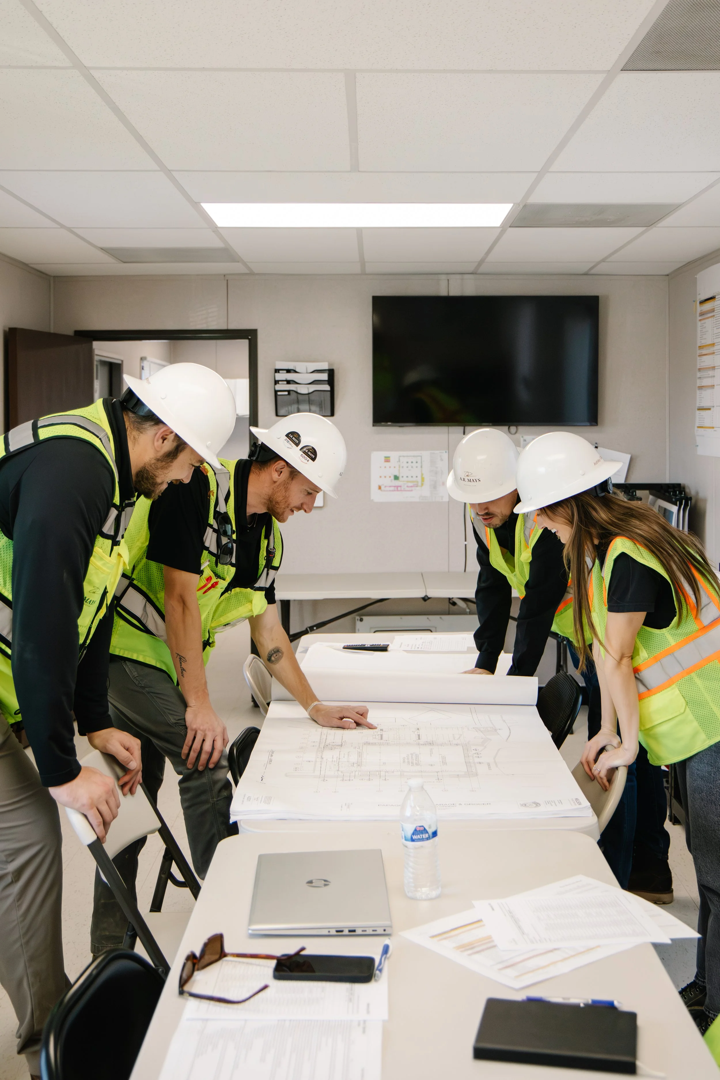 Group of construction workers wearing safety helmets and vests gathered around a table reviewing blueprints in a jobsite trailer.