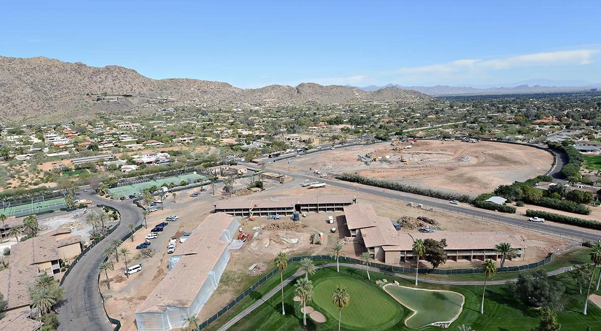 Aerial view of a residential area with a construction site, a small golf course, and mountainous terrain in the background.