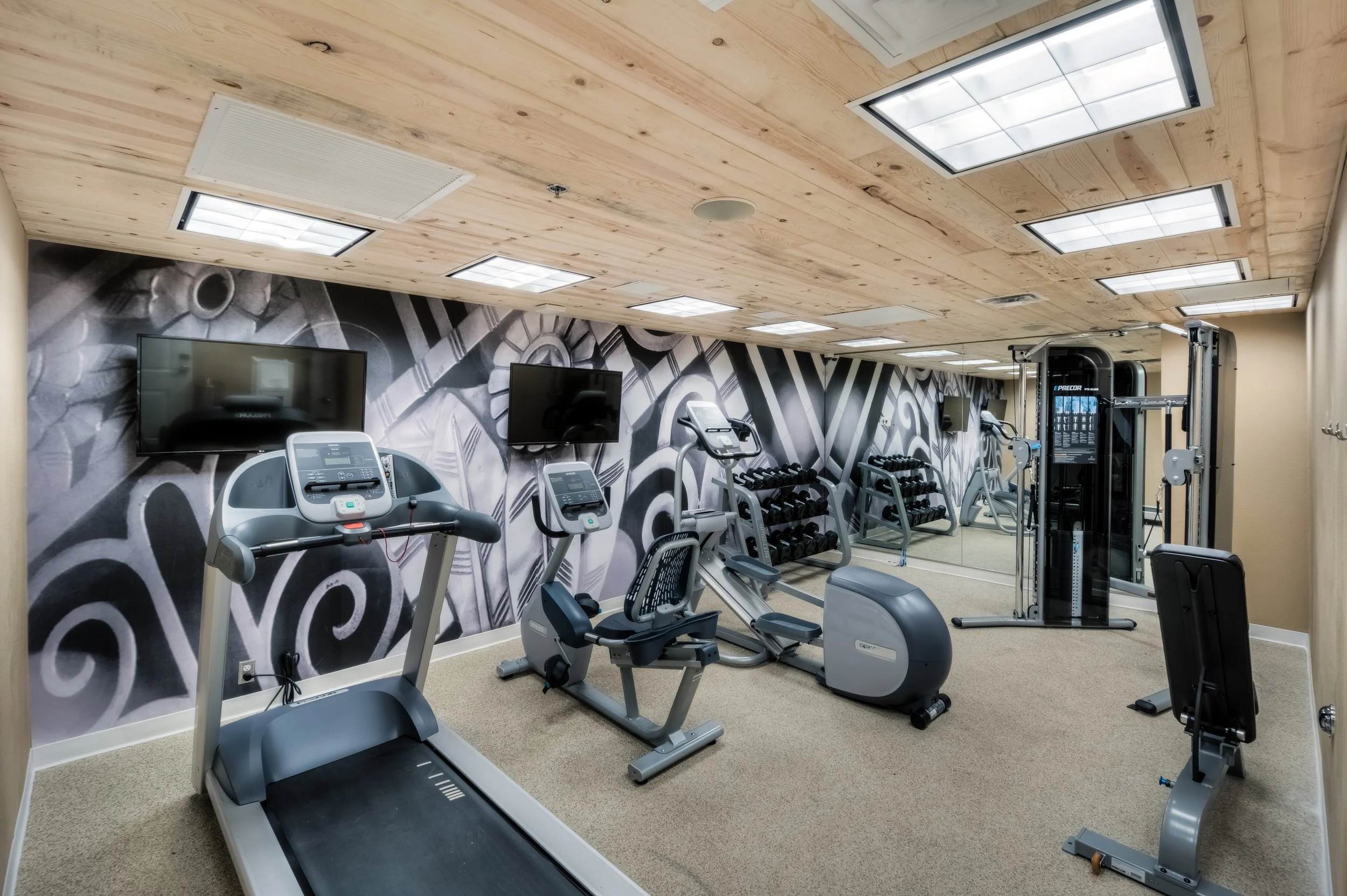 Empty gym room with cardio machines, weight racks, mirrors, and wall-mounted TVs.